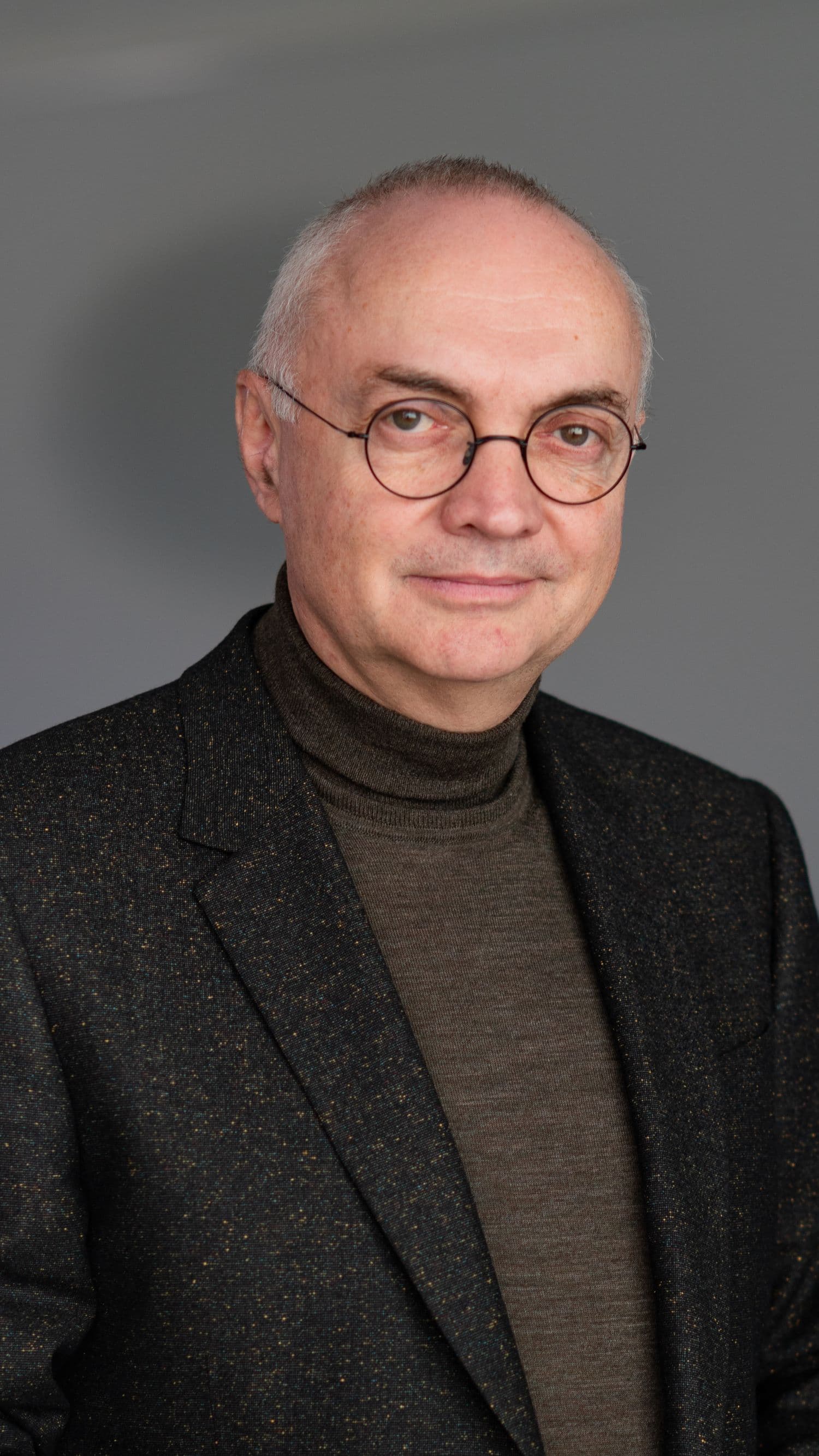 Luc%20Van%20den%20hove_2024_16 Portrait of Luc Van den hove, wearing glasses and a dark blazer against a gray background
