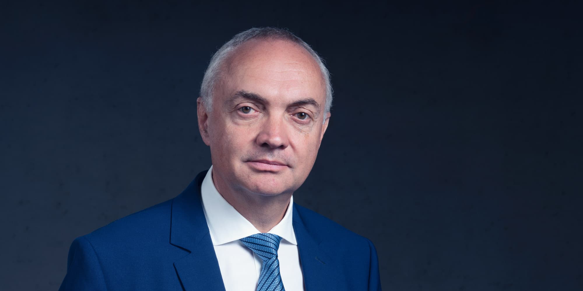 luc-van-den-hove-2023 Portret van Luc Van den hove in een blauw pak tegen een donkere achtergrond