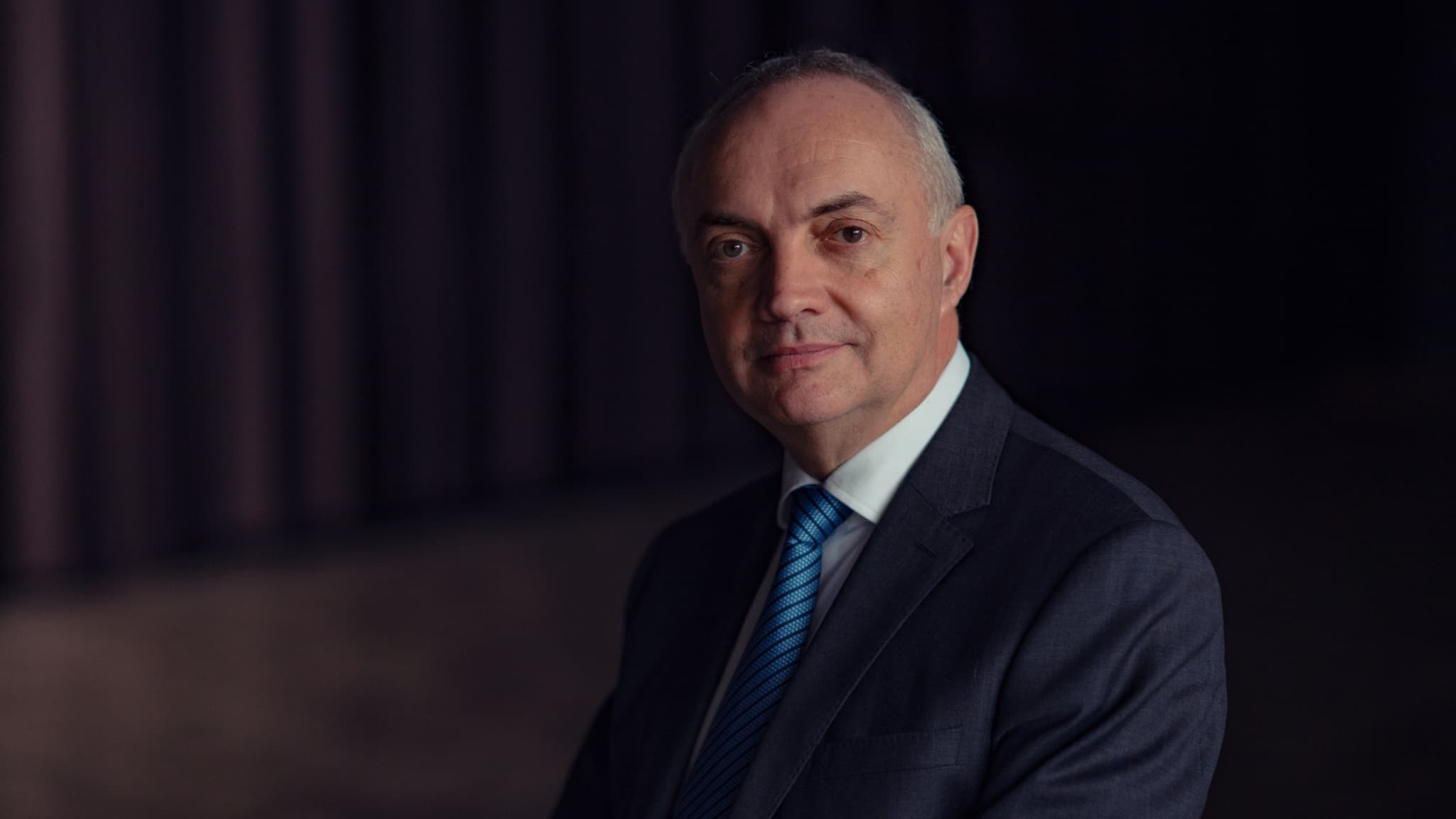 Luc-Van-den-hove-_maxWidth_2048_maxHeight_1152_ppi_72_quality_100_embedMetadata_true_contentETag_6 Portret van een man in een donker pak en blauwe stropdas tegen een donkere achtergrond