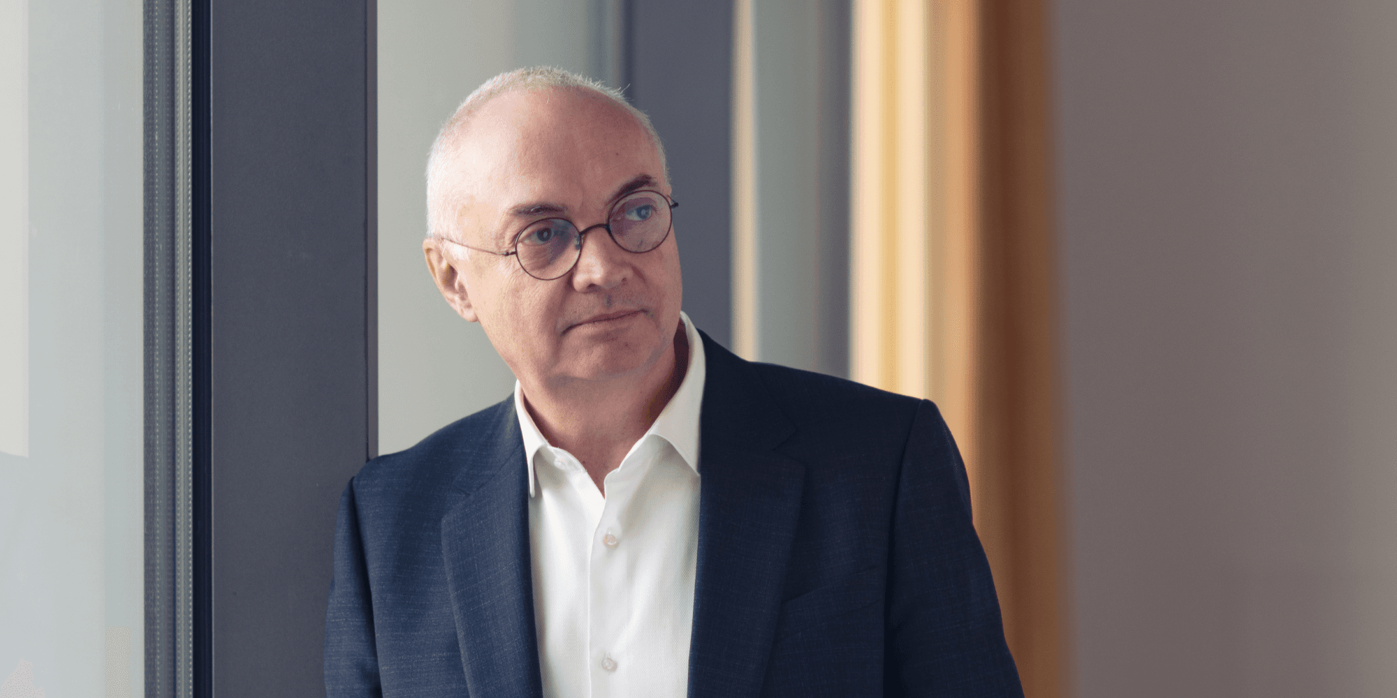 LucVandenhove-1%20%281%29 Luc Van den hove in a dark suit and glasses, standing by a window and looking to the side
