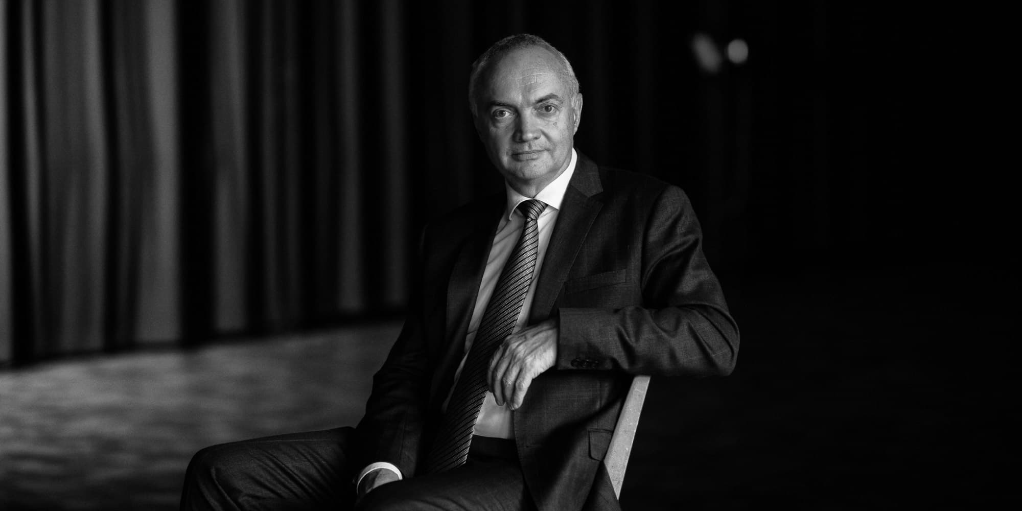lucvandenhove Zwart-witportret van Luc Van den hove, zittend in een pak en stropdas tegen een donkere achtergrond.