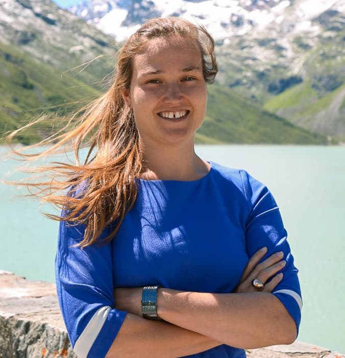 Margot%20Deruyck-2 Margot Deruyck smiling outdoors in a blue top with mountains and a lake behind her