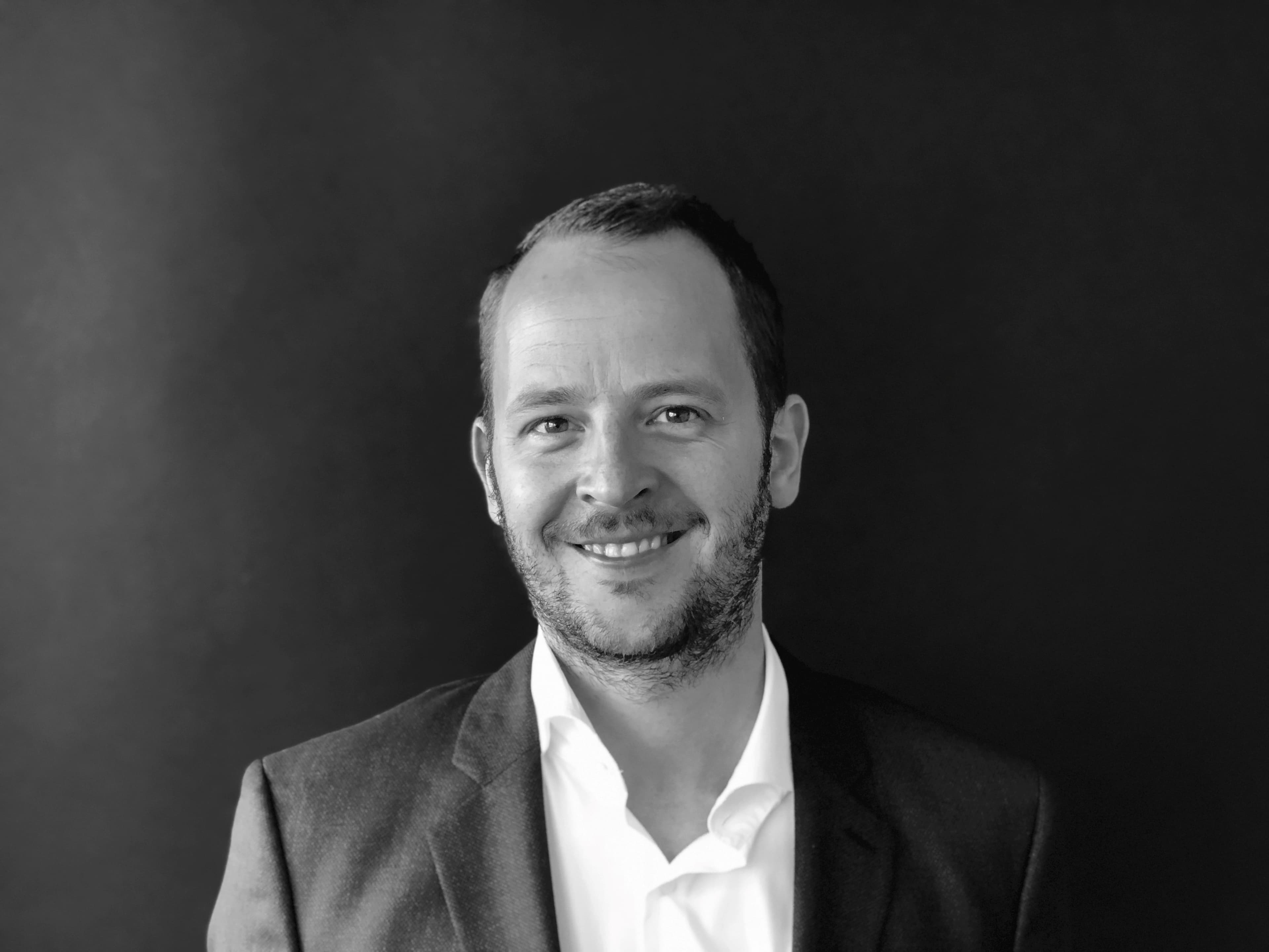 MartijnJoris_1 Zwart-witportret van Martijn Joris in een blazer en wit overhemd, glimlachend tegen een donkere achtergrond.