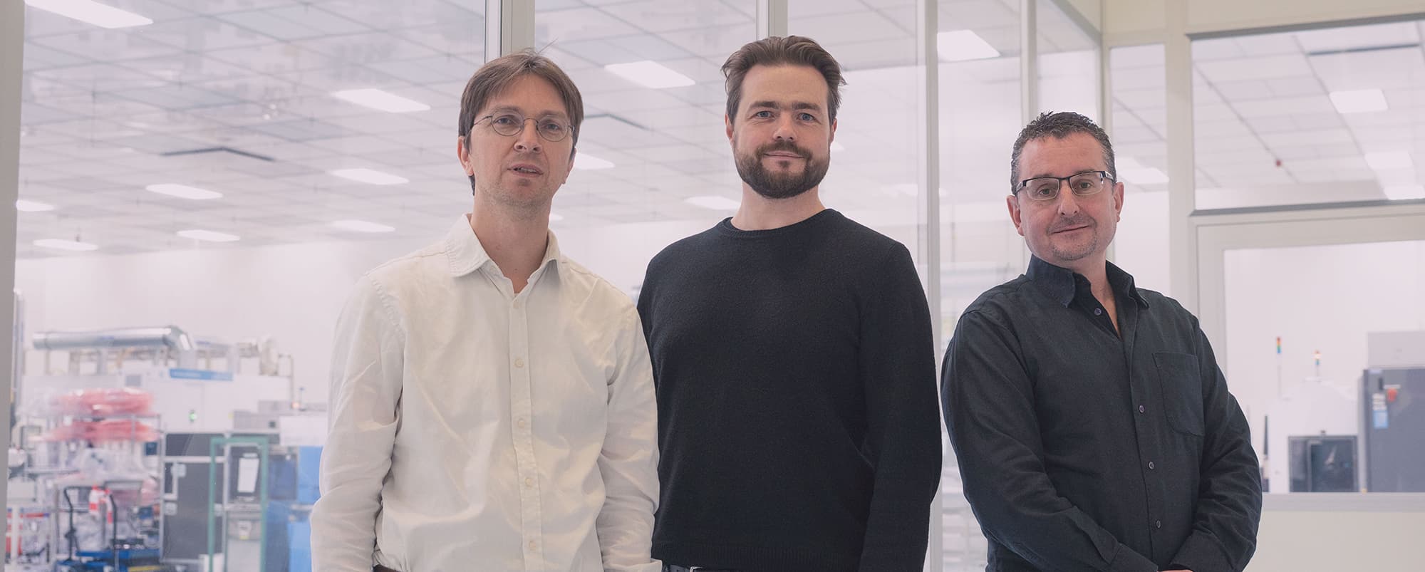 micledi-team002_3 Drie mannen staan in een cleanroomlaboratorium, met apparatuur zichtbaar achter glazen wanden