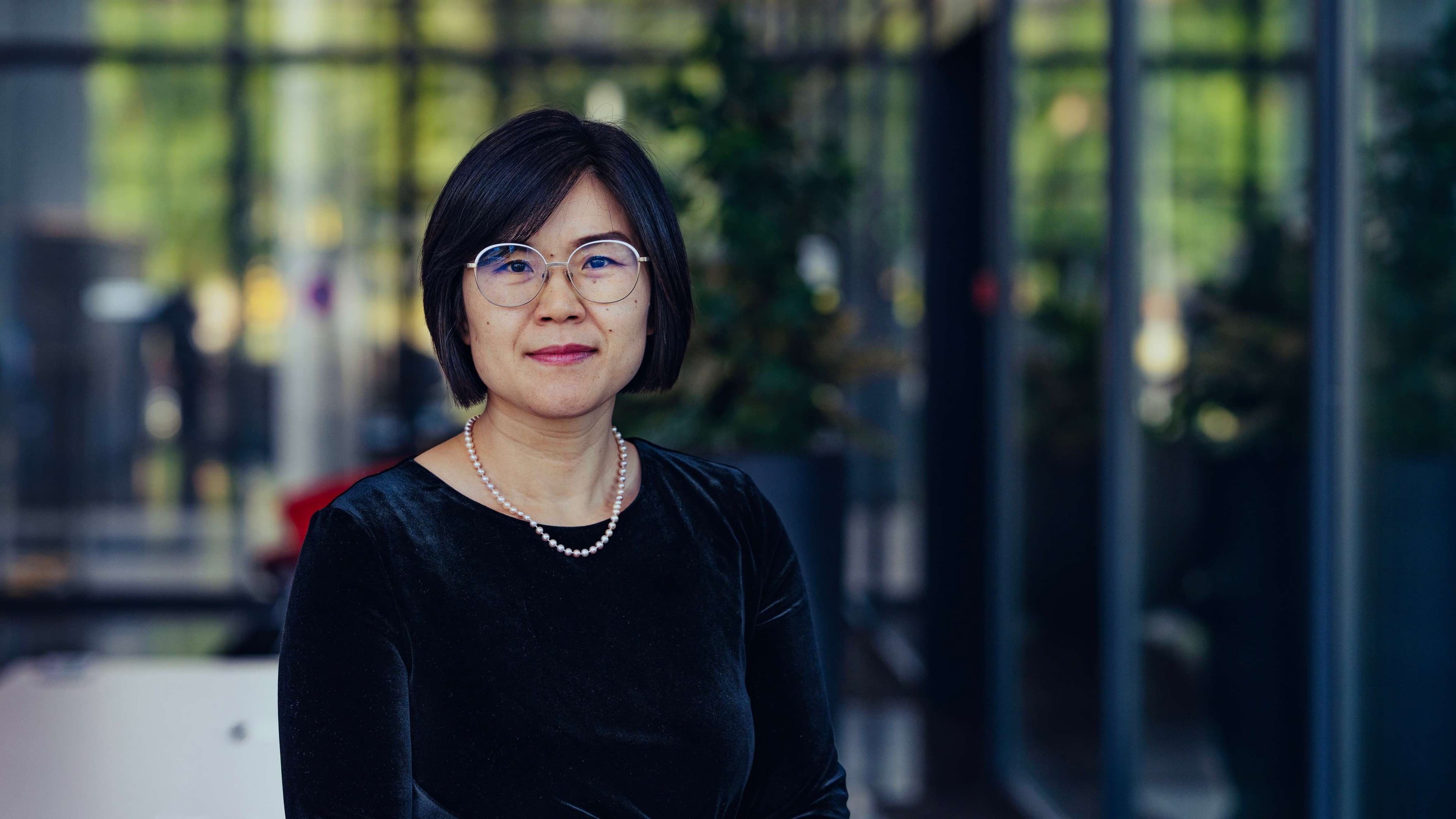 Minghui Portrait of Minghui Huang wearing glasses and a black top, standing indoors against a blurred glass background.