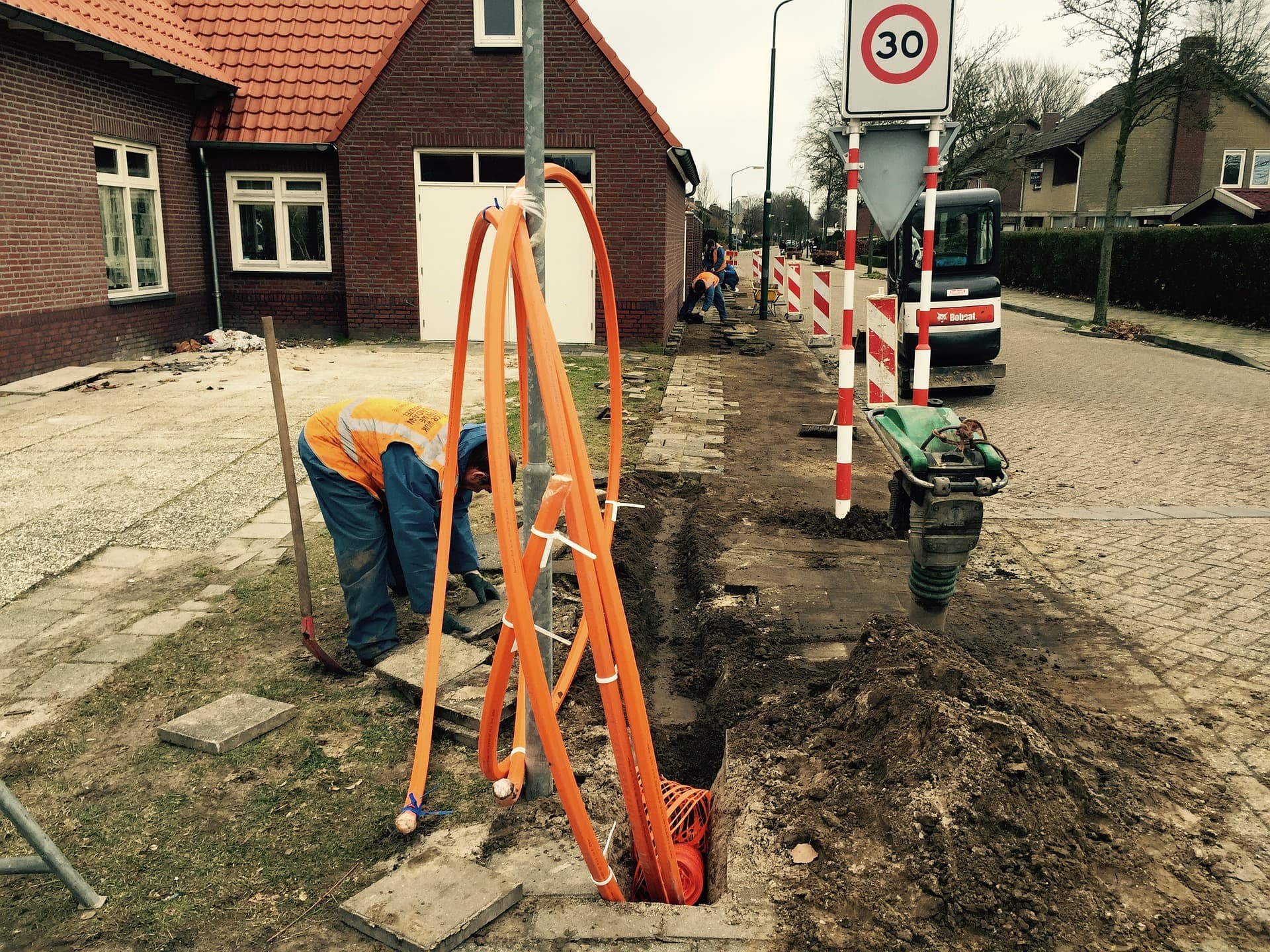 MM-WAVES_Header%20Image Werknemers die oranje glasvezelbuis leggen in een sleuf in een woonstraat naast een 30 km/u-bord.