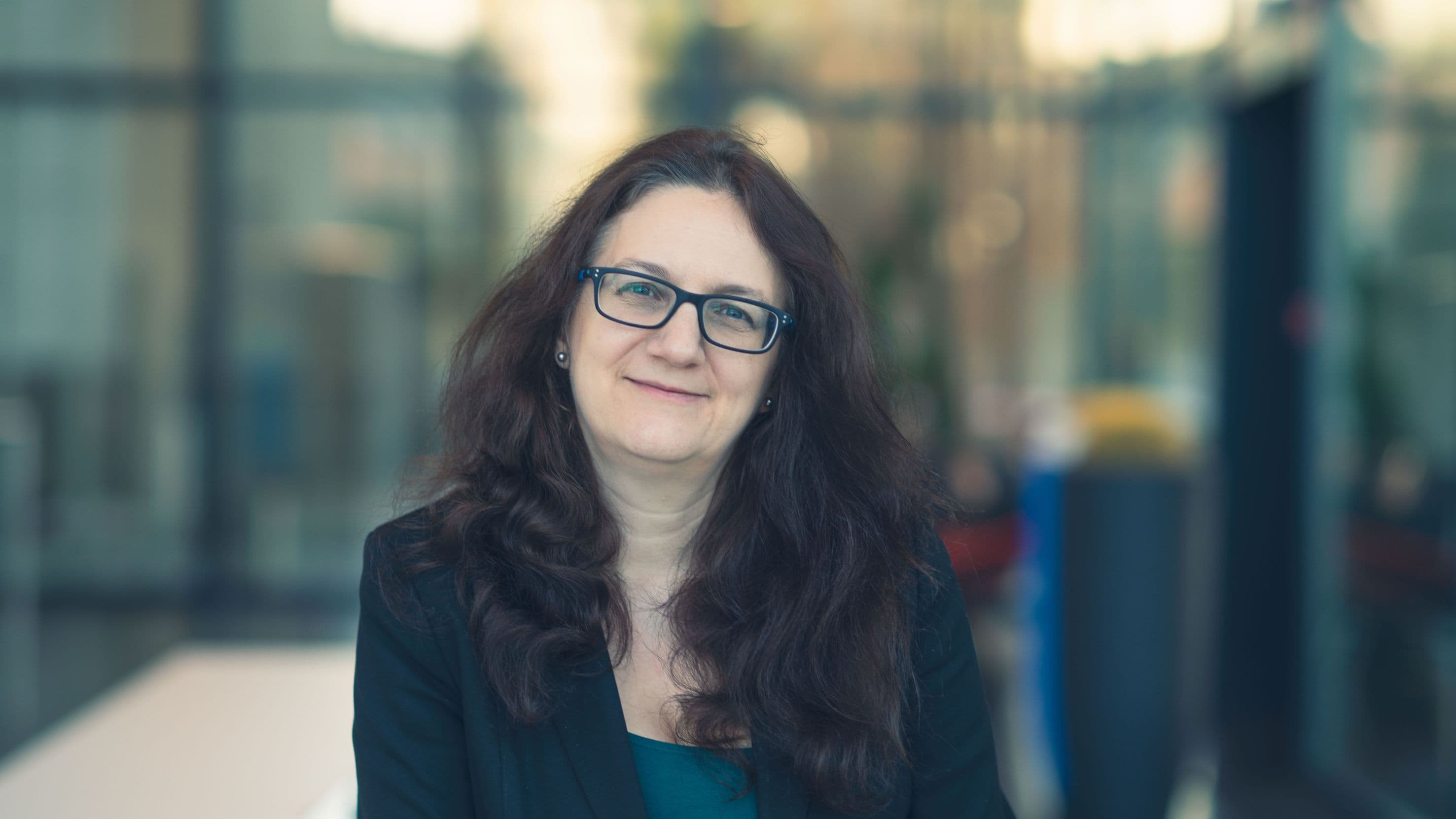 nadine_collaert Portrait of Nadine Collaert with long dark hair and glasses, standing in a softly blurred indoor setting.