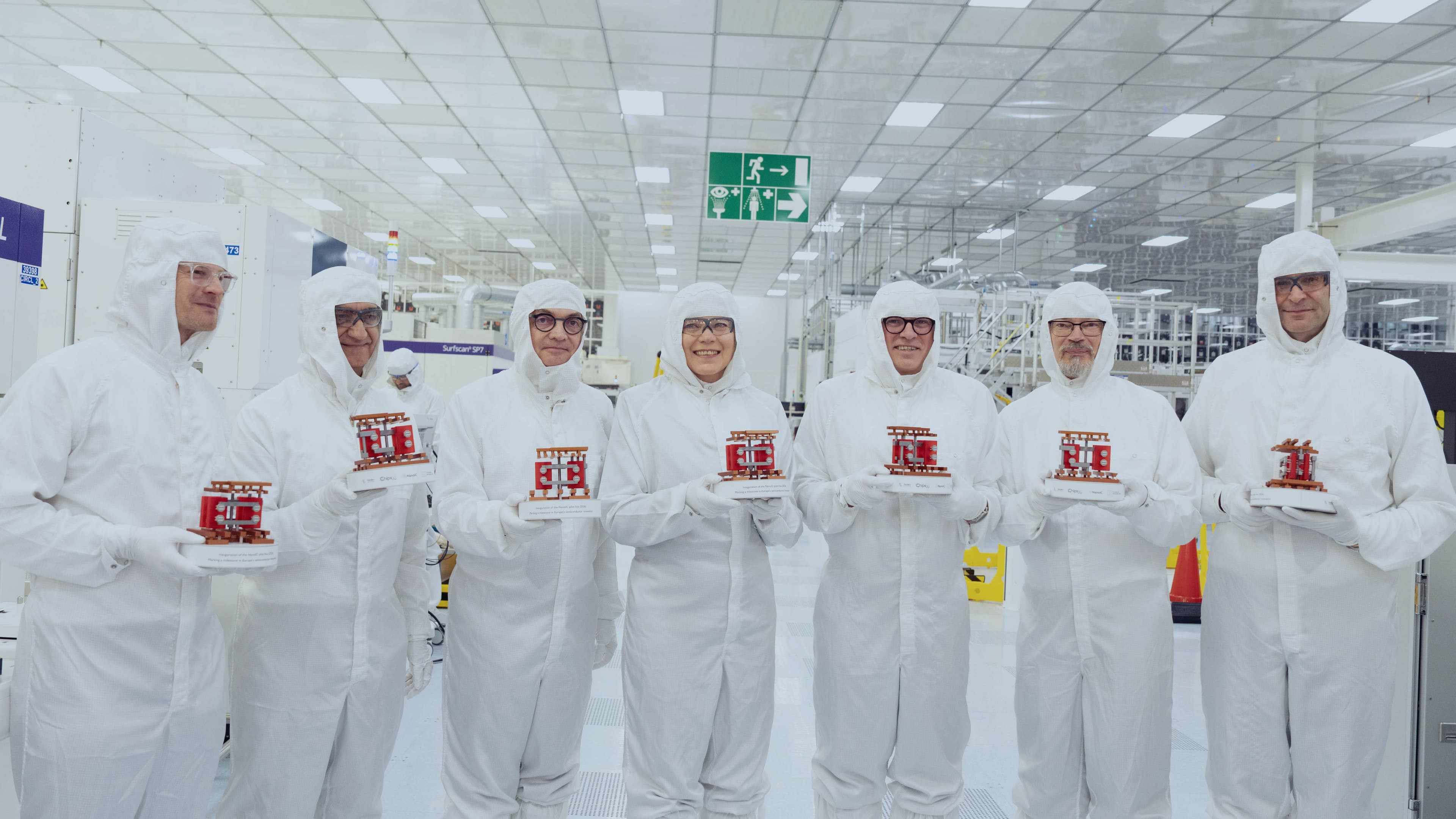 NANOIC2_header Seven people in cleanroom suits pose in a semiconductor cleanroom, each holding a small NanoIC model.