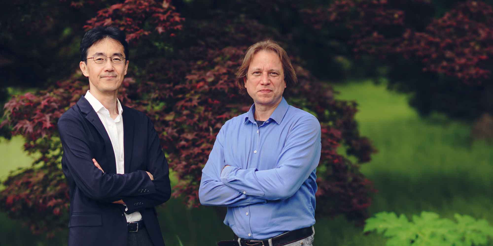 naoto-zsolt-imec Two imec researchers stand with arms crossed in front of a blurred leafy background.