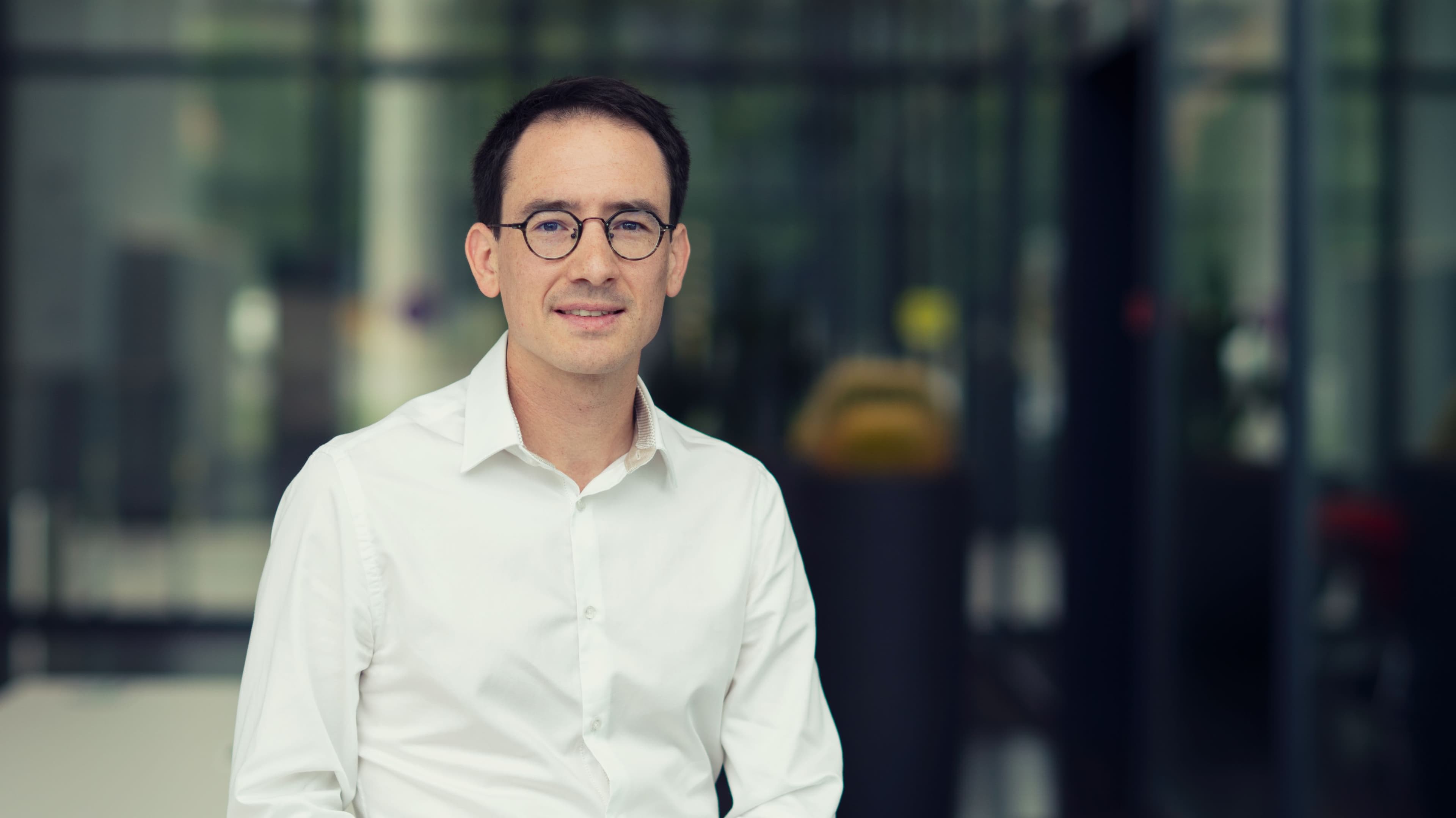 Nicolas%20Pantano Portrait of Nicolas Pantano in a white shirt and glasses, standing in front of a blurred glass building interior