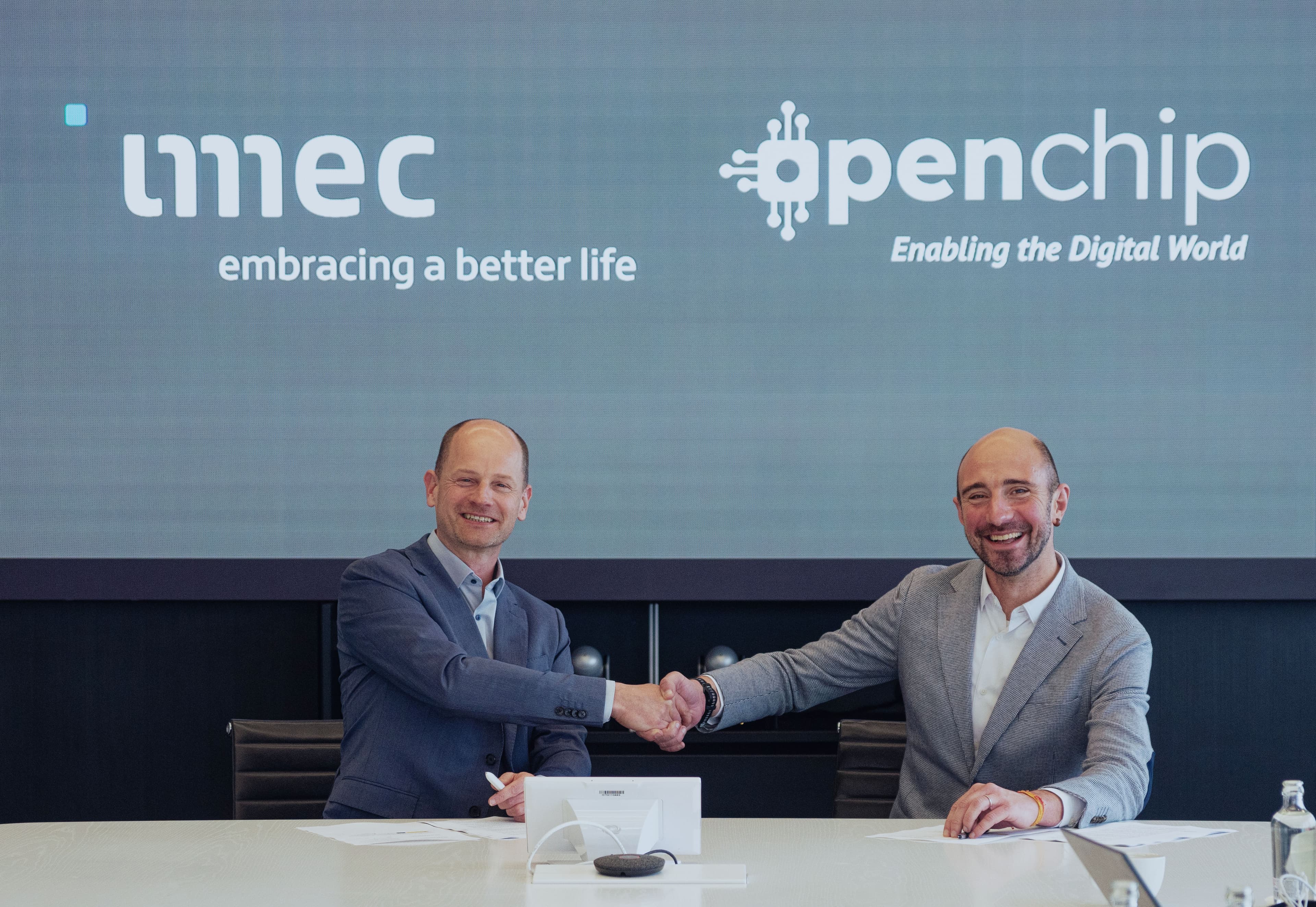 Open%20Chip00004 Patrick Vandenameele and Cesc Guim shake hands in front of imec and Openchip logos.