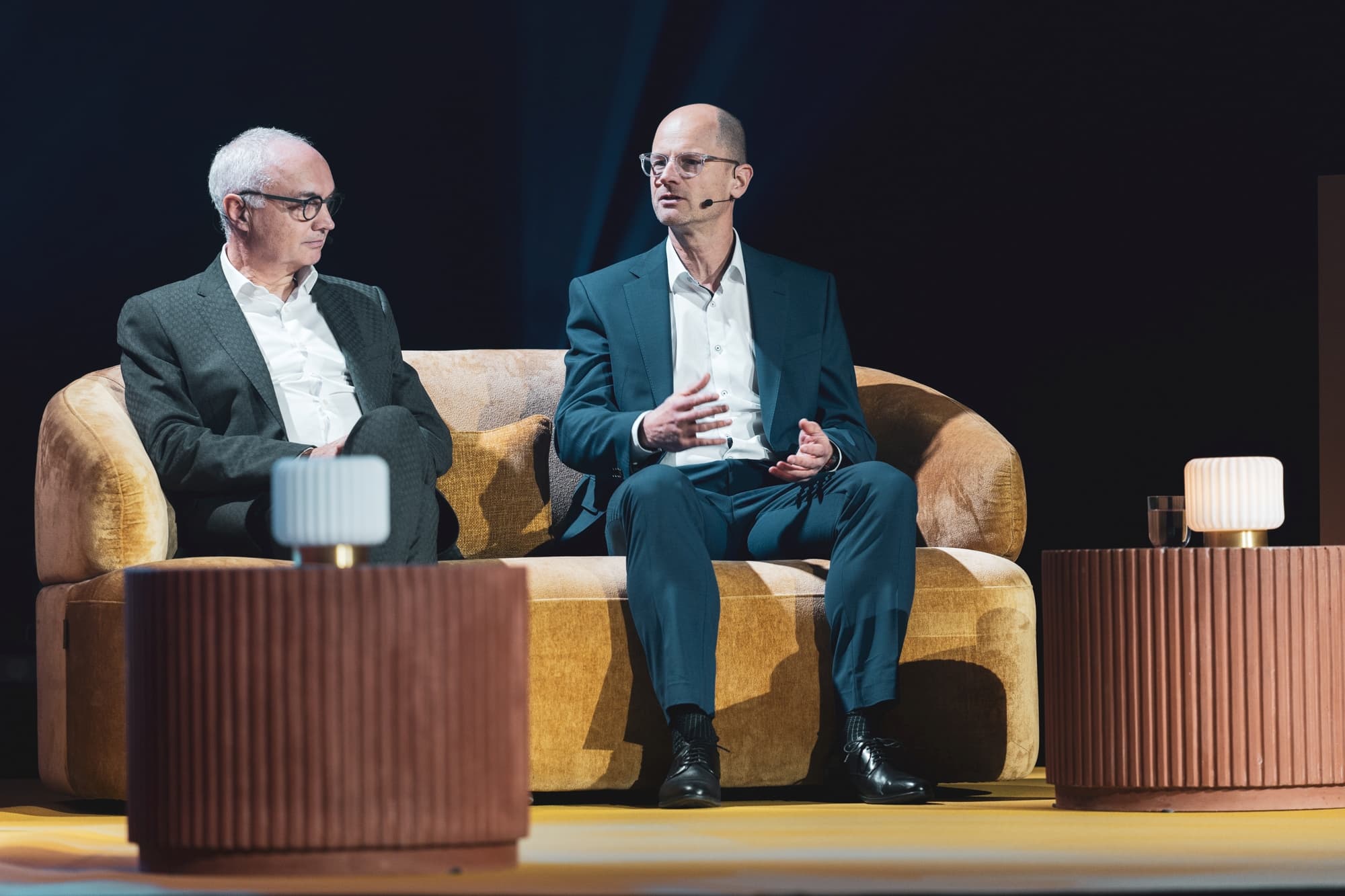 openminds2025-023 Twee mannen in pakken die op het podium spreken tijdens een paneldiscussie, zittend op een beige bank met microfoons.