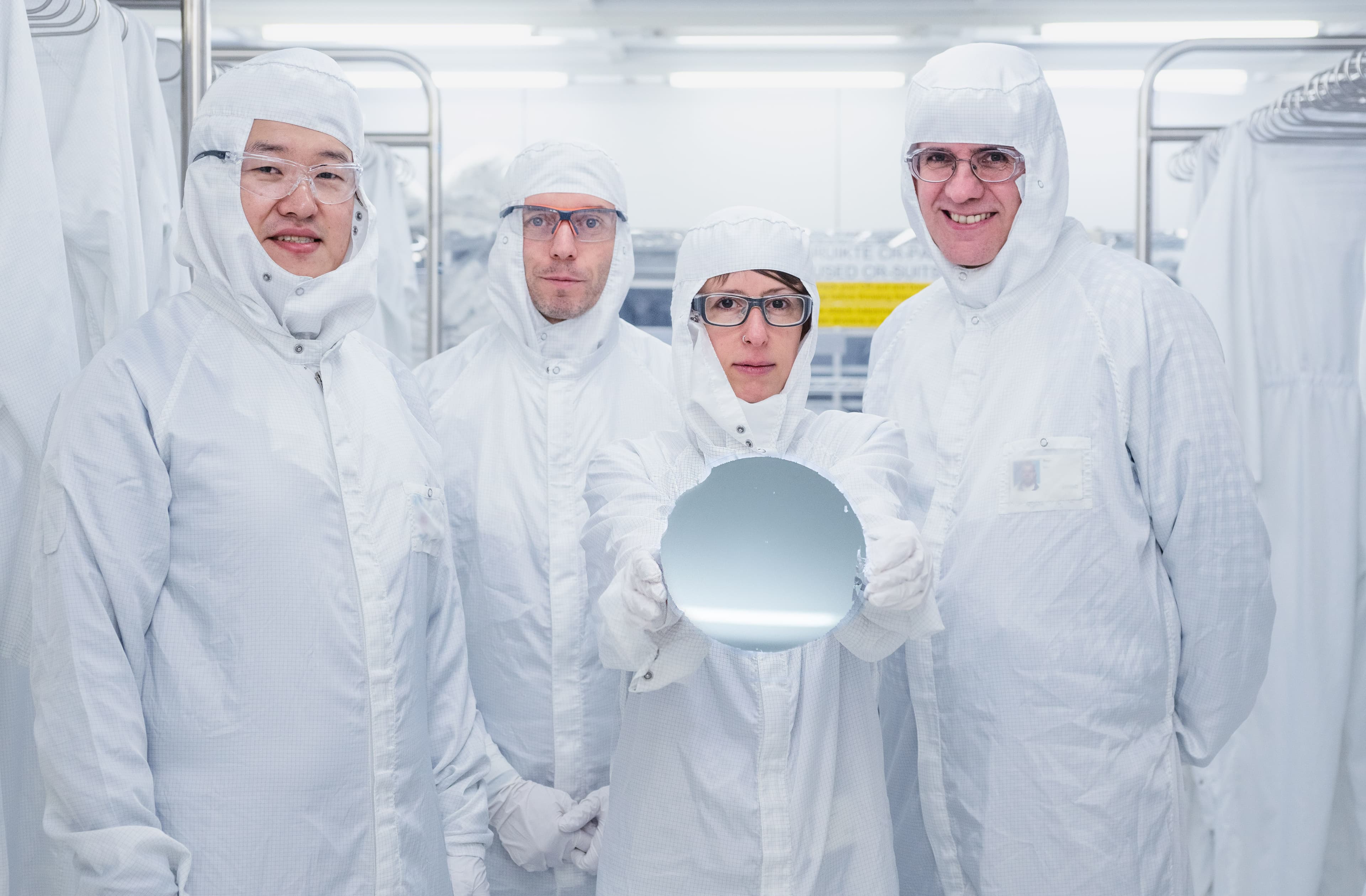 Photo%201 Onderzoekers in cleanroompakken houden een reflecterende wafer vast in een cleanroom