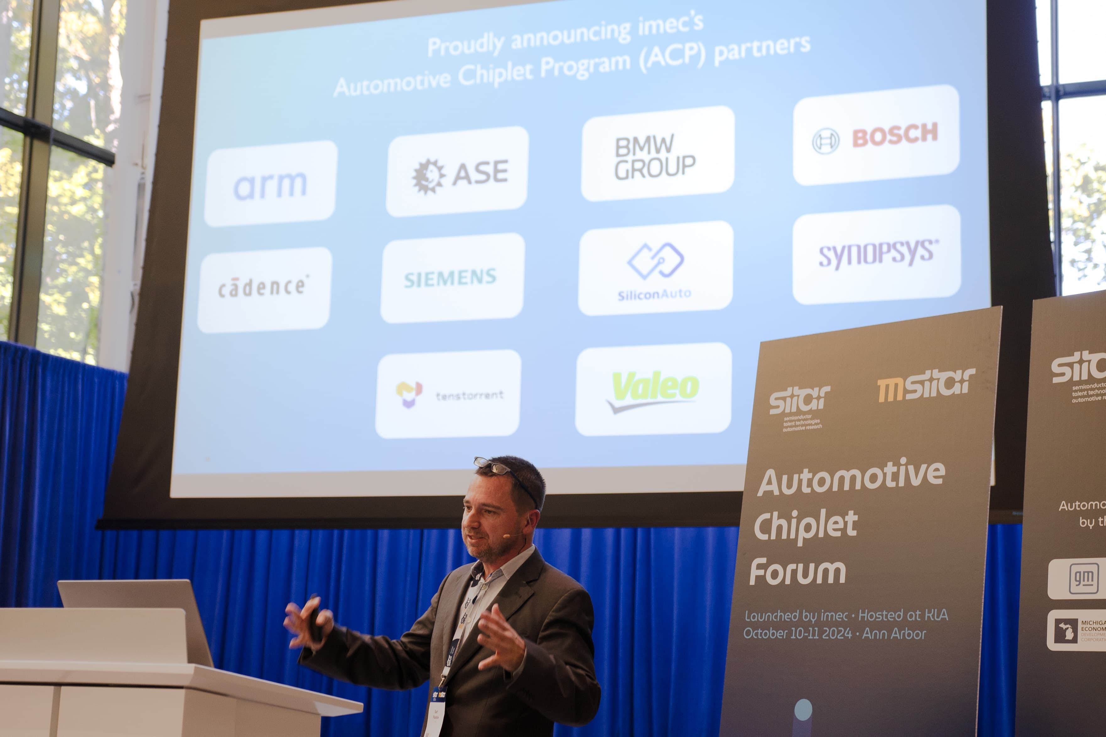 Speaker presenting Automotive Chiplet Program with partner logos Speaker presenting the Automotive Chiplet Program with partner logos on a projection screen
