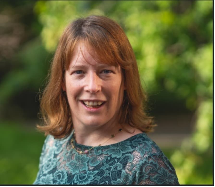 Portrait einer Frau mit türkisfarbenem Oberteil im Freien Liesbet Lagae smiling in a teal patterned top against a blurred green outdoor background.