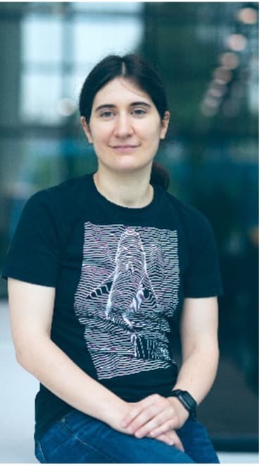 Portrait von Charlotte Herzeel vor unscharfem Hintergrund Charlotte Herzeel seated in front of a blurred hallway, wearing a black graphic T-shirt.