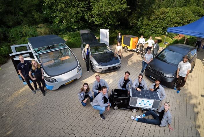 Group beside solar-powered cars and prototype vehicle Group posing beside several solar-equipped cars and a small prototype vehicle at an outdoor event