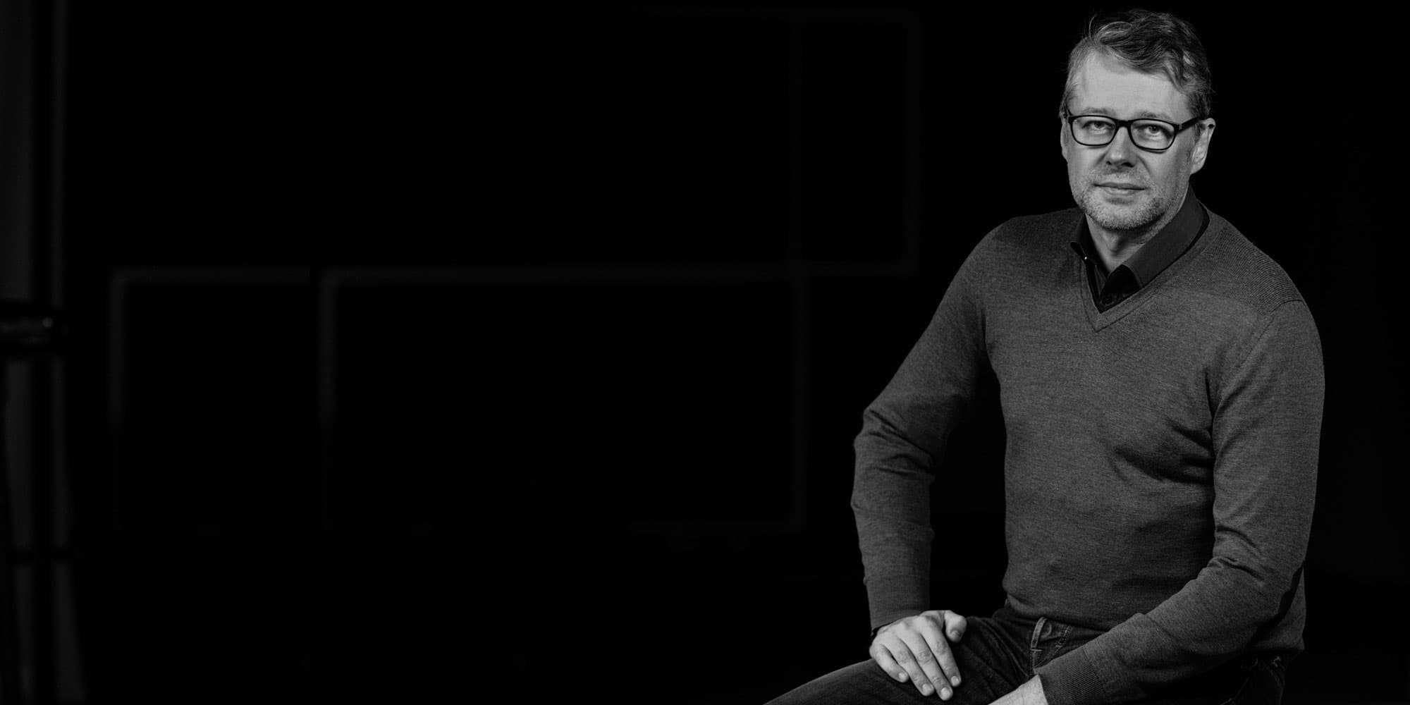 pieter-ballon002magazinenew_1 Black-and-white portrait of Pieter Ballon seated against a dark background, wearing glasses and a sweater.