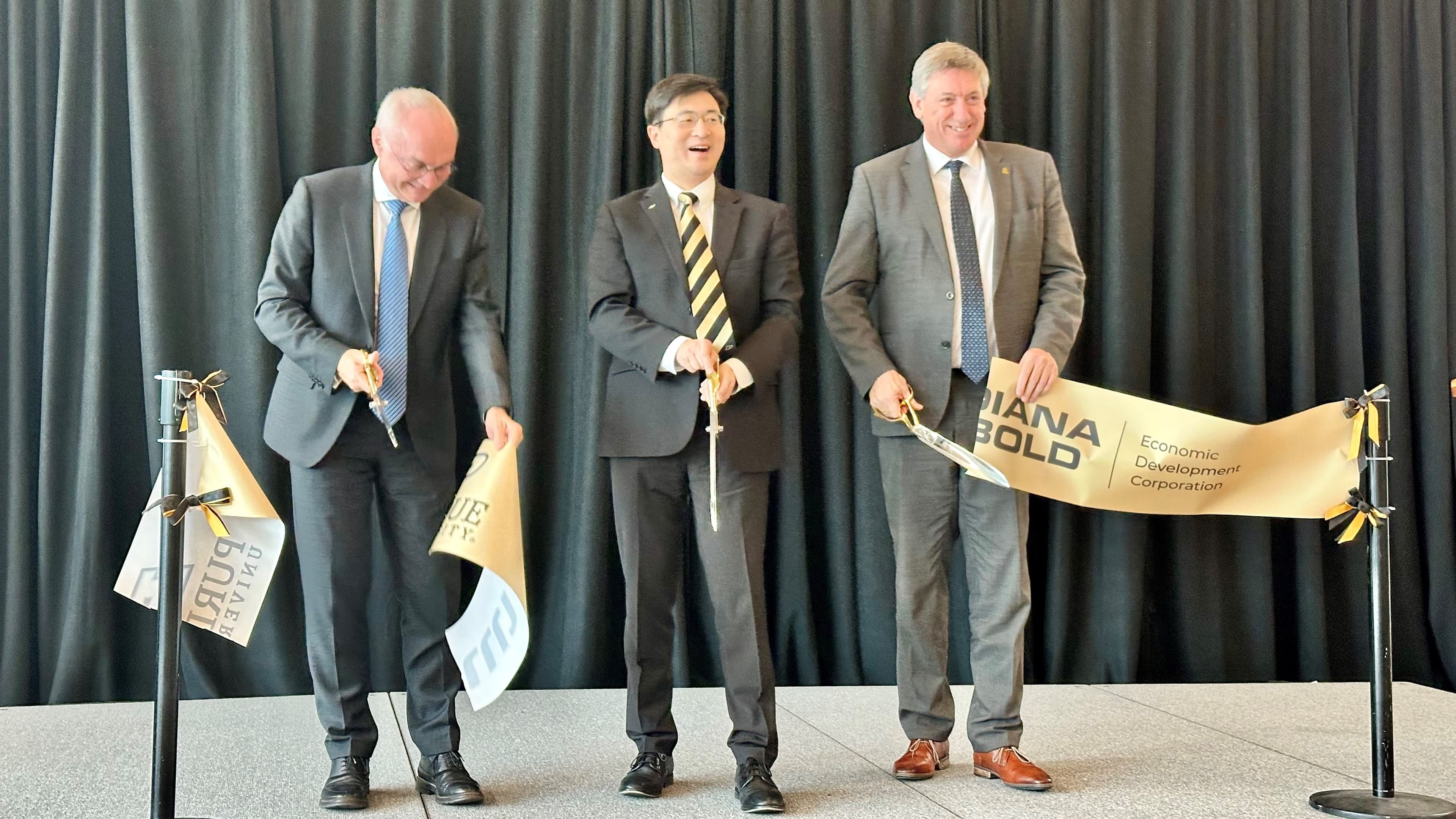 Purdue_2 Three men in suits cut a ceremonial ribbon at a Purdue event stage.
