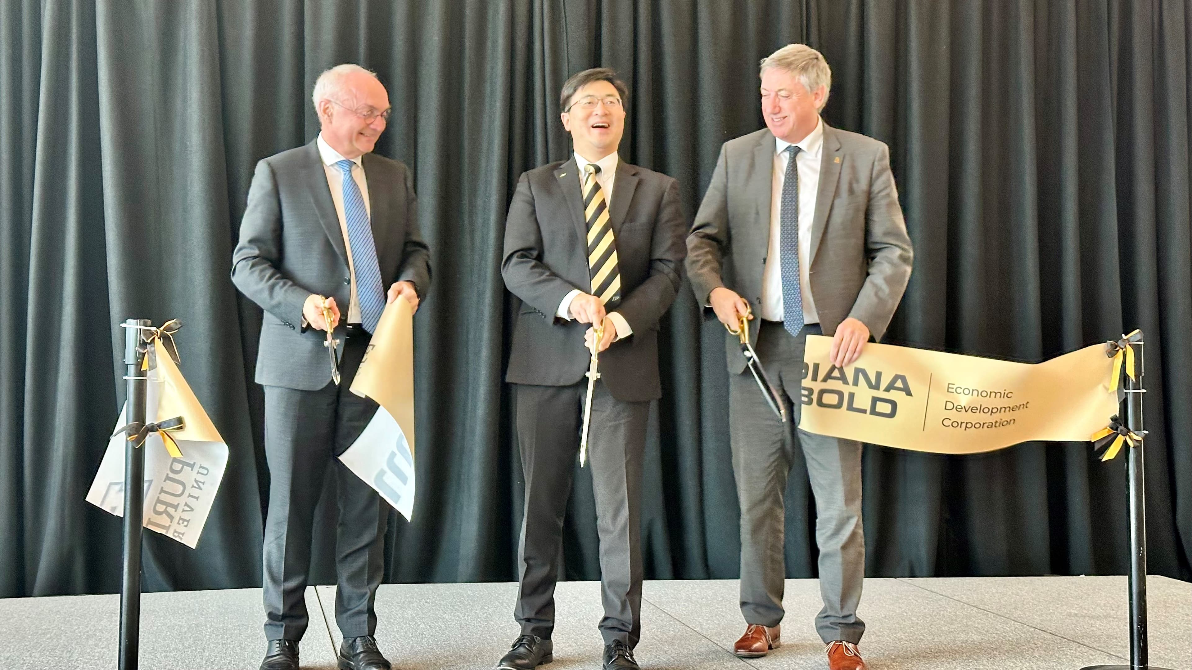 Purdue Three suited men cut a ribbon at the Purdue–Indiana Bold event, standing before black curtains.