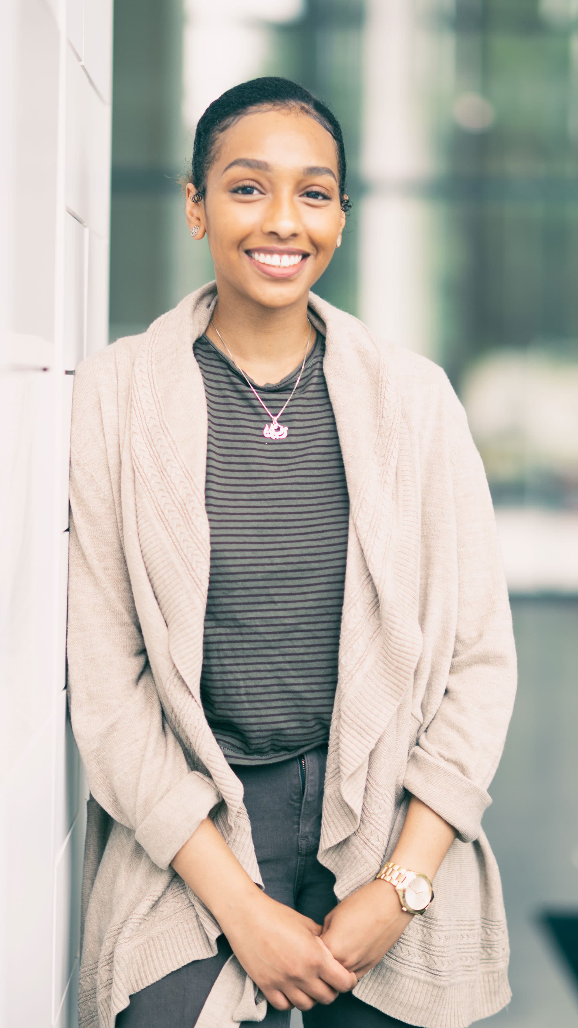 Rayyan_1 Smiling woman standing indoors beside a glass wall, wearing a beige cardigan and striped shirt.