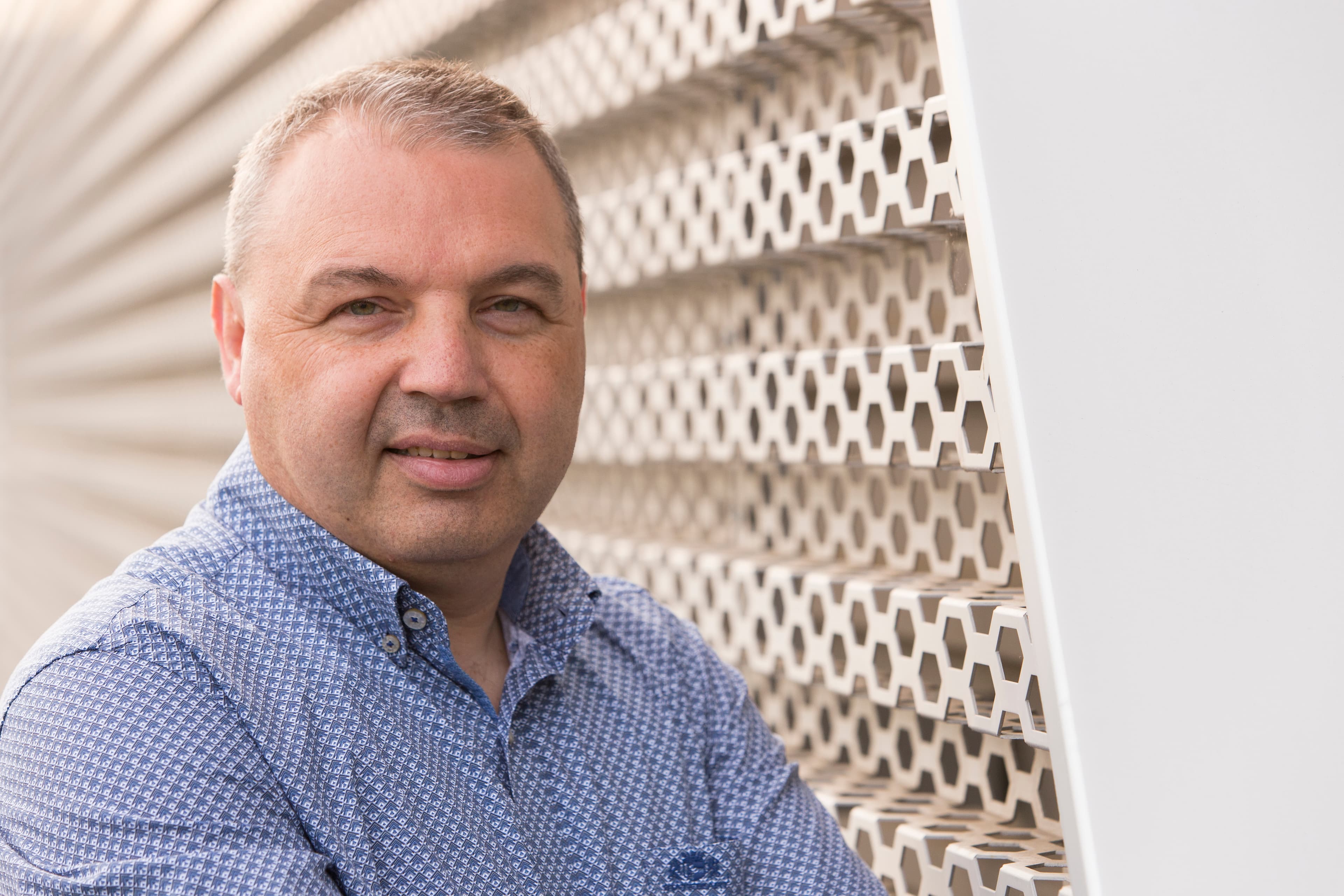 Ronny_Vrijens_1 Portrait of Ronny Vrijens in a patterned blue shirt beside a white honeycomb wall