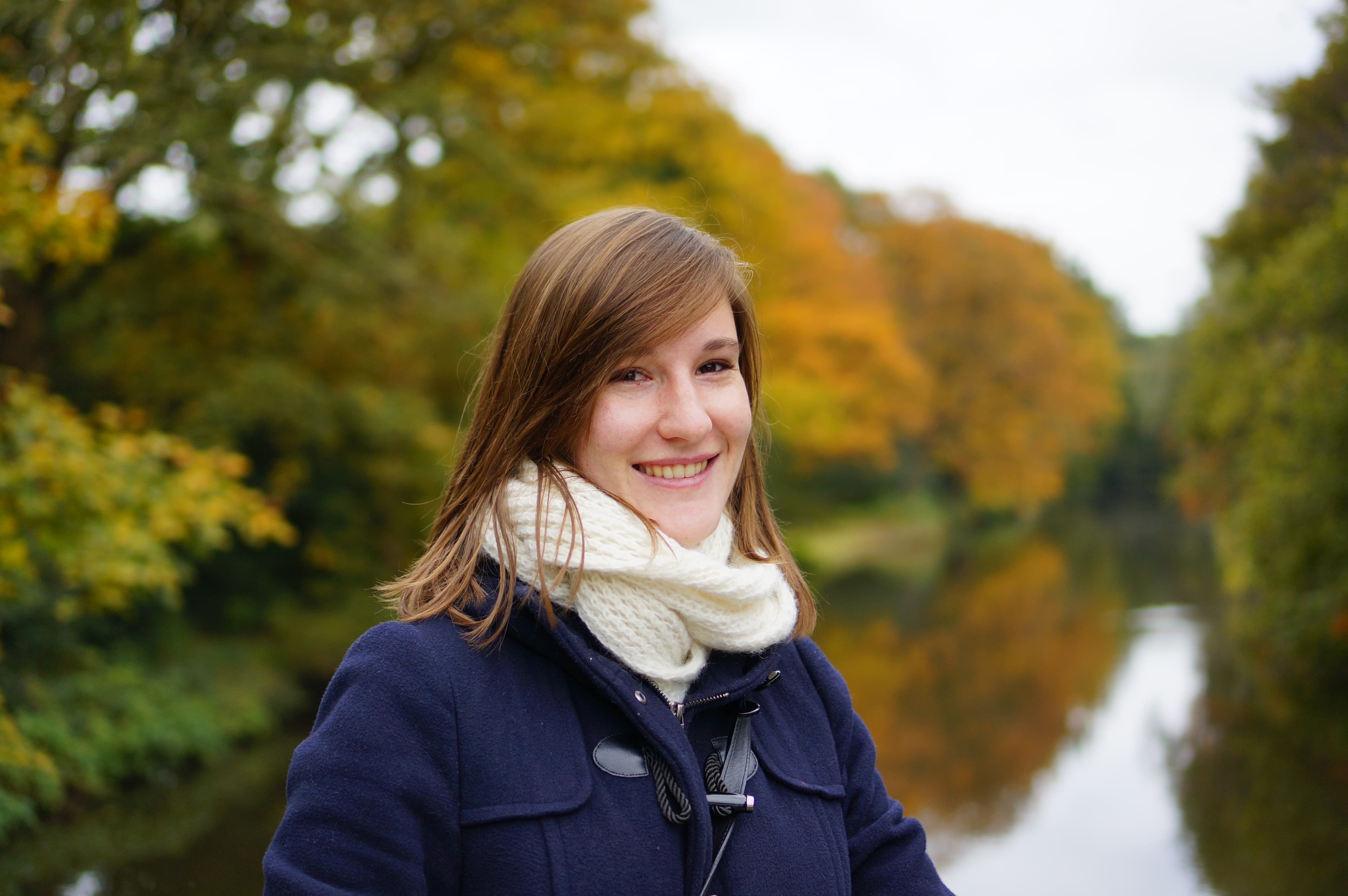 sarah__1 Smiling woman in a navy coat and white scarf standing by a canal with autumn trees behind her