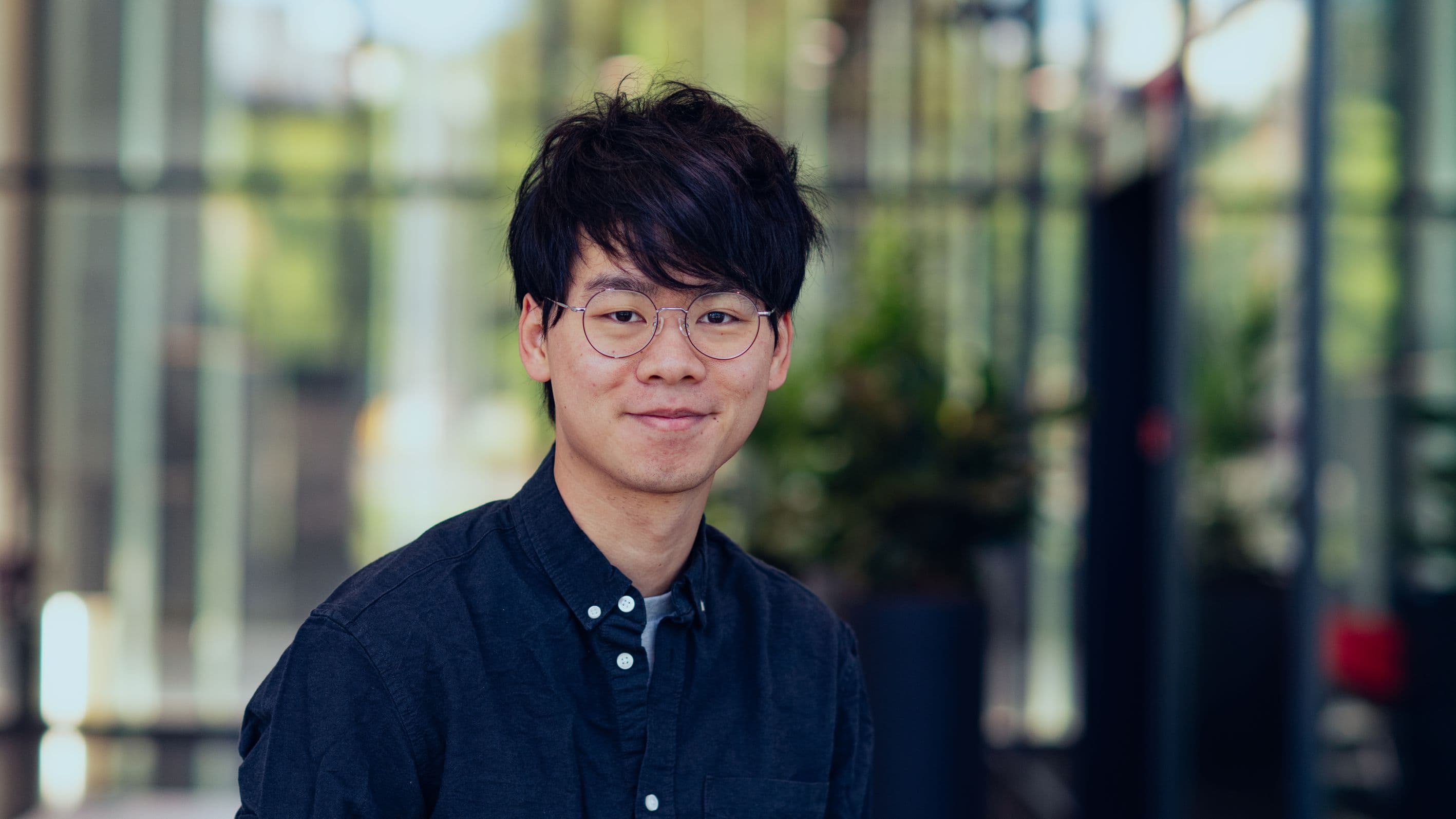 Shane%20Lin Portrait of Shane Lin wearing glasses and a dark shirt, standing in front of a blurred glass building
