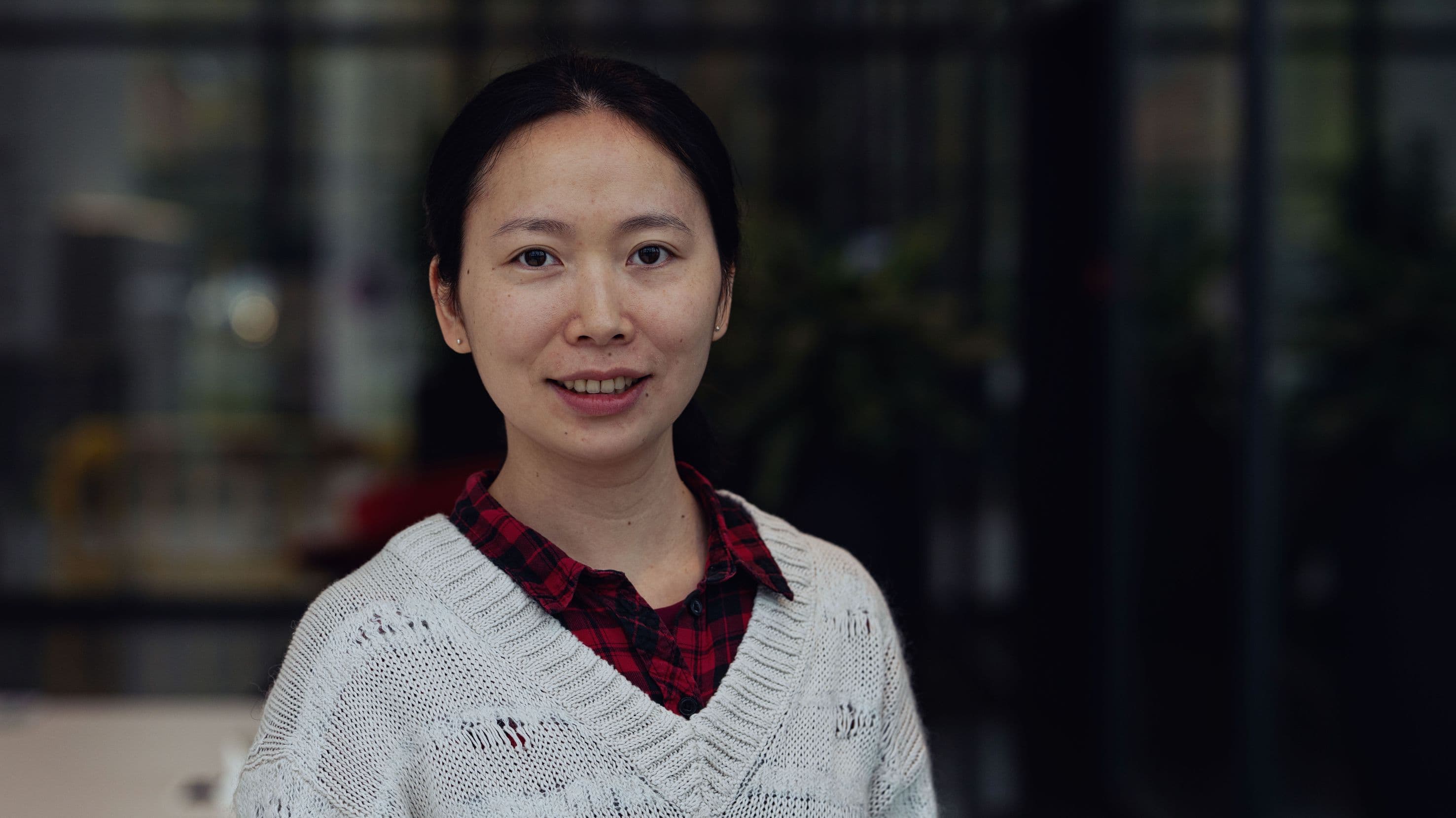 Sheng%20Yang Portrait of researcher Sheng Yang in a white sweater and red plaid shirt, standing against a blurred lab background.