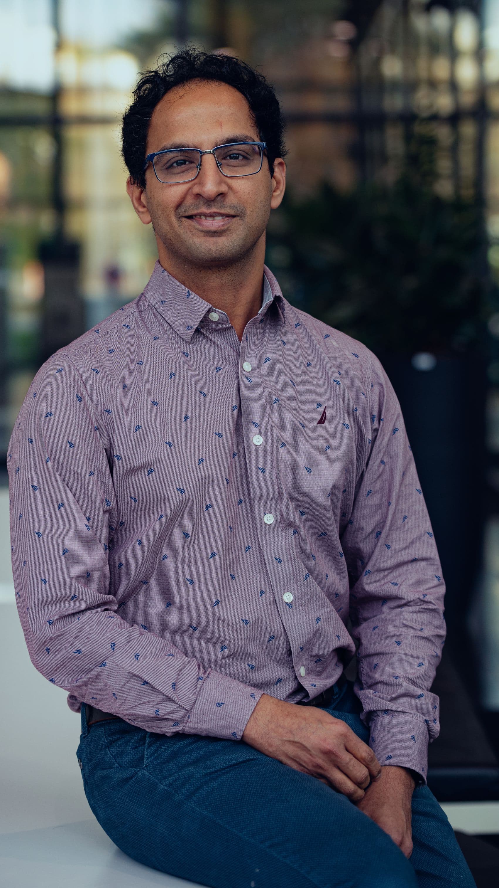 Siddharth%20Rao Portrait of Siddharth Rao, wearing glasses and a patterned shirt, seated indoors and smiling at the camera.