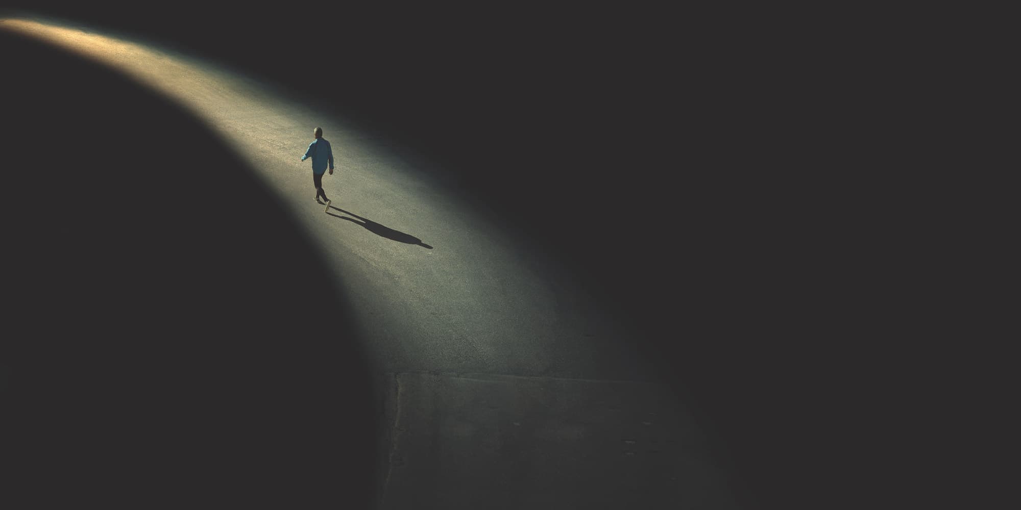 Lone pedestrian under spotlight on dark street A lone pedestrian walks through a dark street lit by a single bright beam of light.