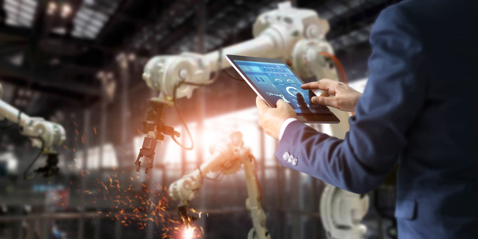 smartproductinnovation_1 Person in a suit using a tablet to monitor industrial robotic arms in a factory