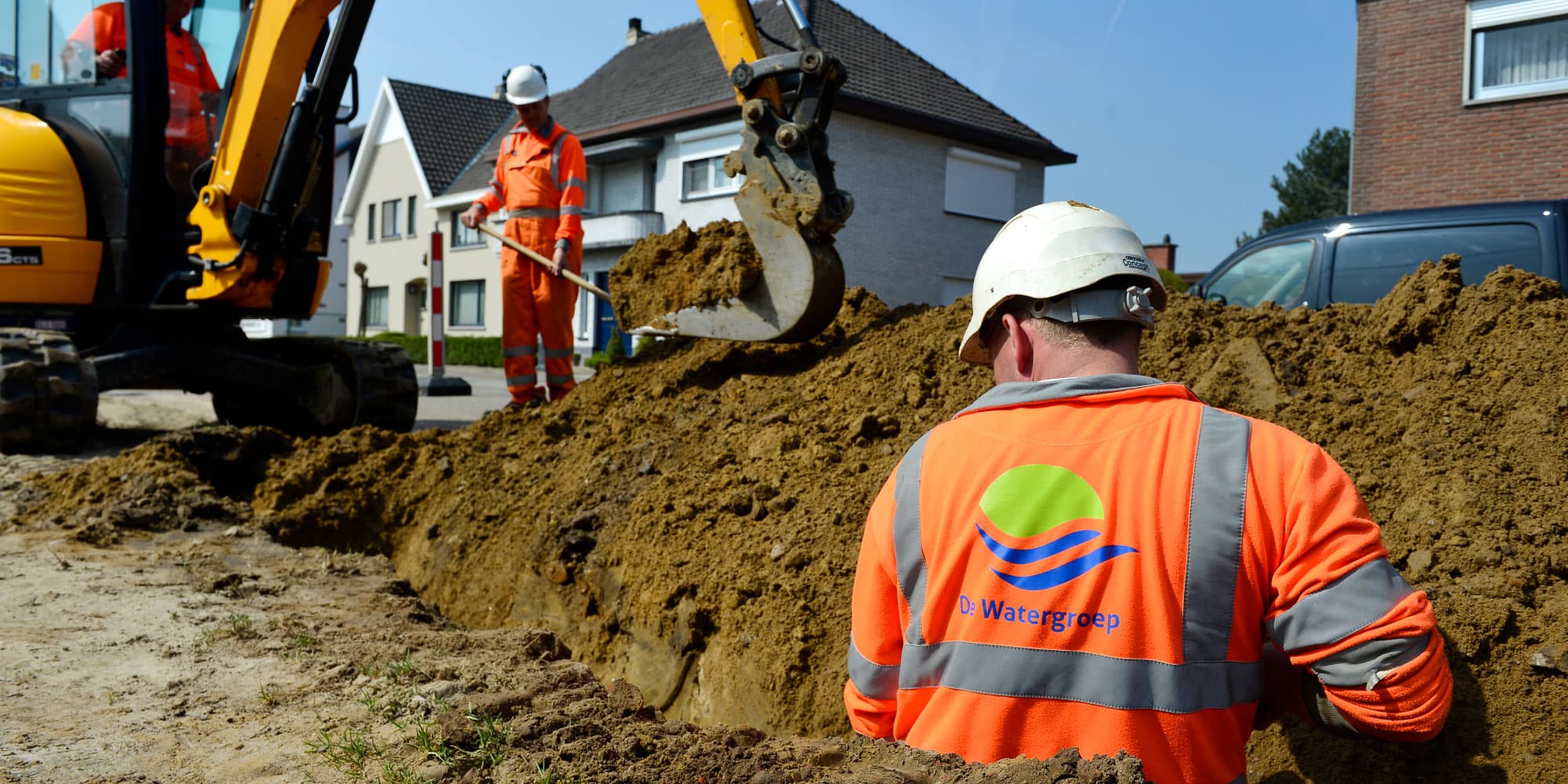 smartwatergrid Nutswerkers in oranje veiligheidskleding graven een sleuf met een graafmachine in een woonstraat.