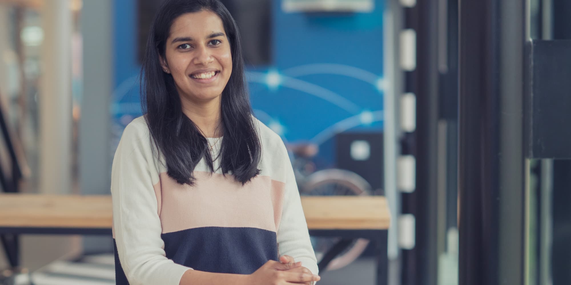 Sneha Portrait of Sneha, smiling indoors in front of a blue research-themed background