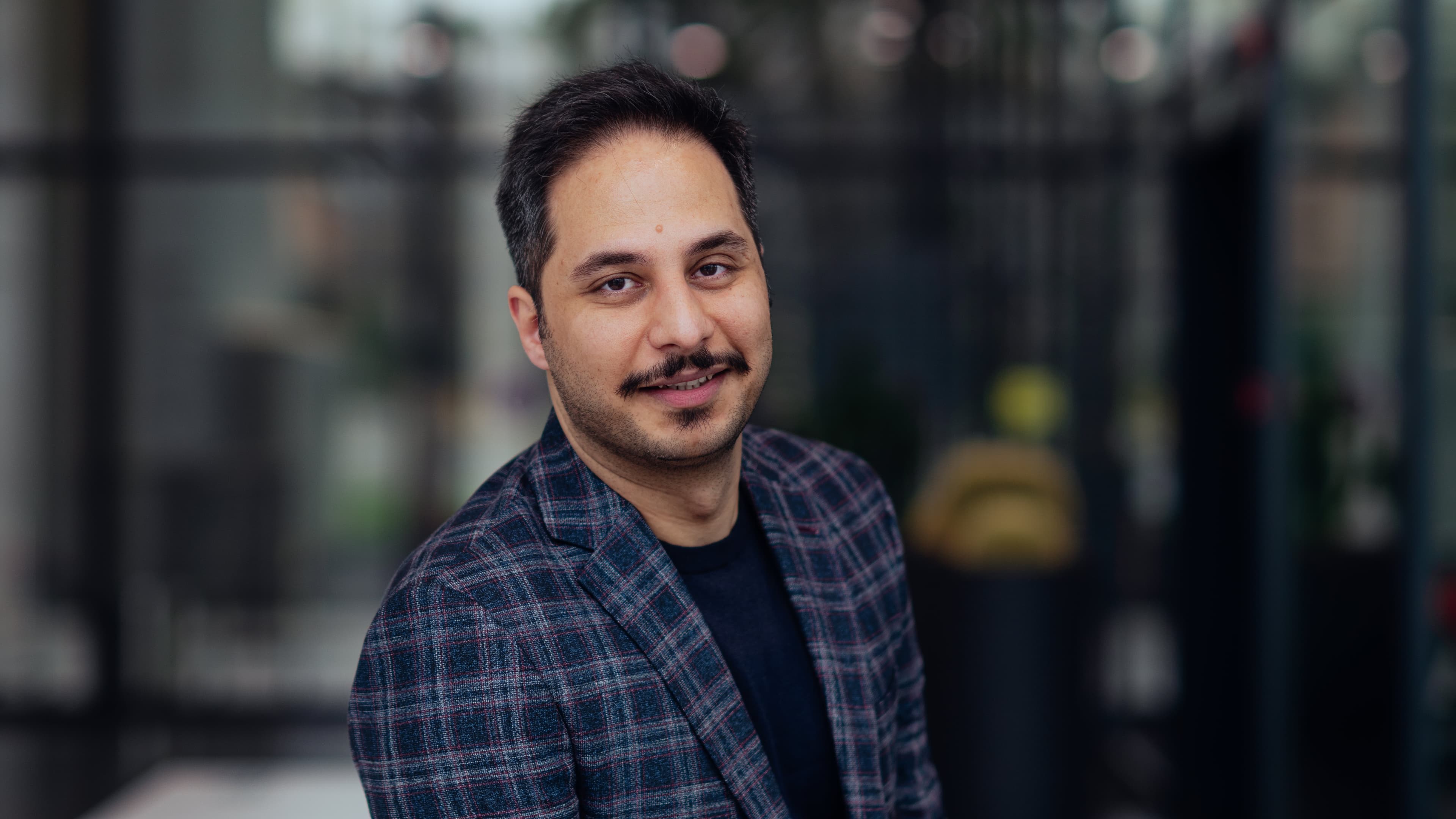 Sohrab-Pahlavan001-min Portrait of a smiling man in a blazer, standing in front of a blurred lab or office background.