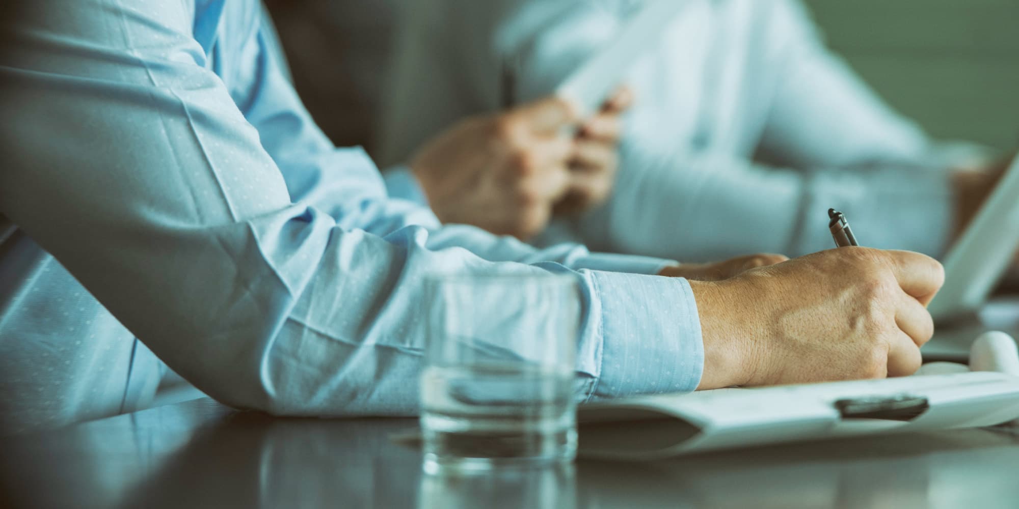 startup-article Two people in light blue shirts take notes during a meeting, with a glass and table in the foreground.