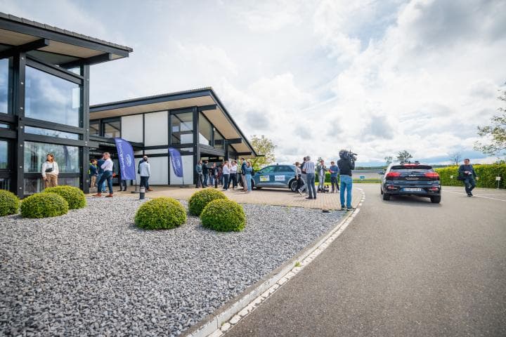 Testweek%20telebediend%20rijden%20in%20Lommel Mensen verzamelen zich buiten een modern gebouw naast twee geparkeerde auto's tijdens een test van teleoperatief rijden in Lommel.