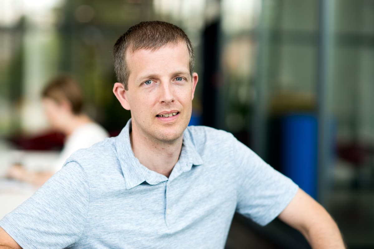 Tom_Torfs_RS_1 Portrait of Tom Torfs in a light blue polo shirt, seated indoors with a blurred colleague in the background.