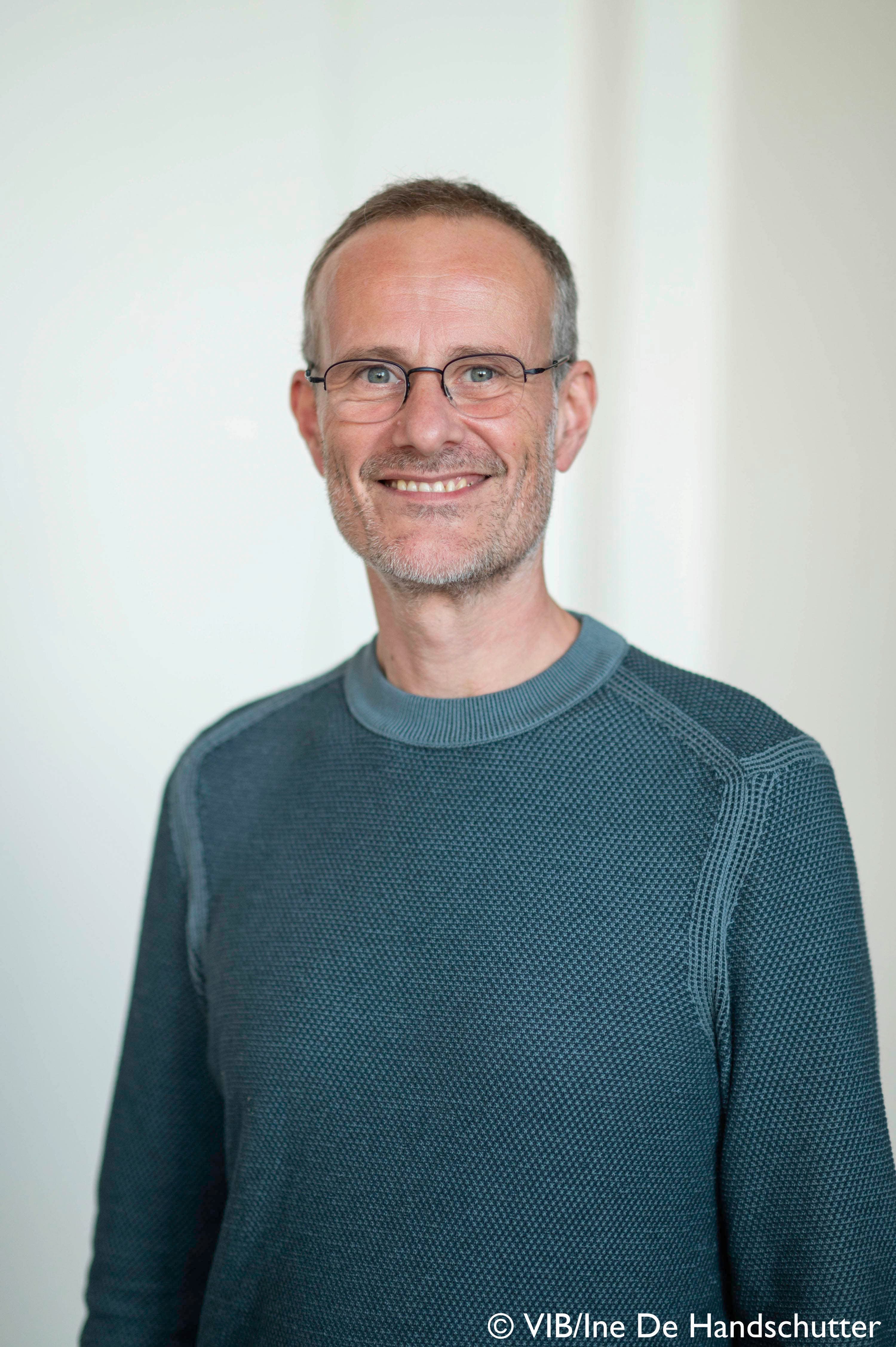 vib-Ine%20De%20Handschutter%20-%20Patrik%20Verstreken%20%281%29 Portrait of Patrik Verstreken smiling in a dark teal sweater and glasses against a light background.