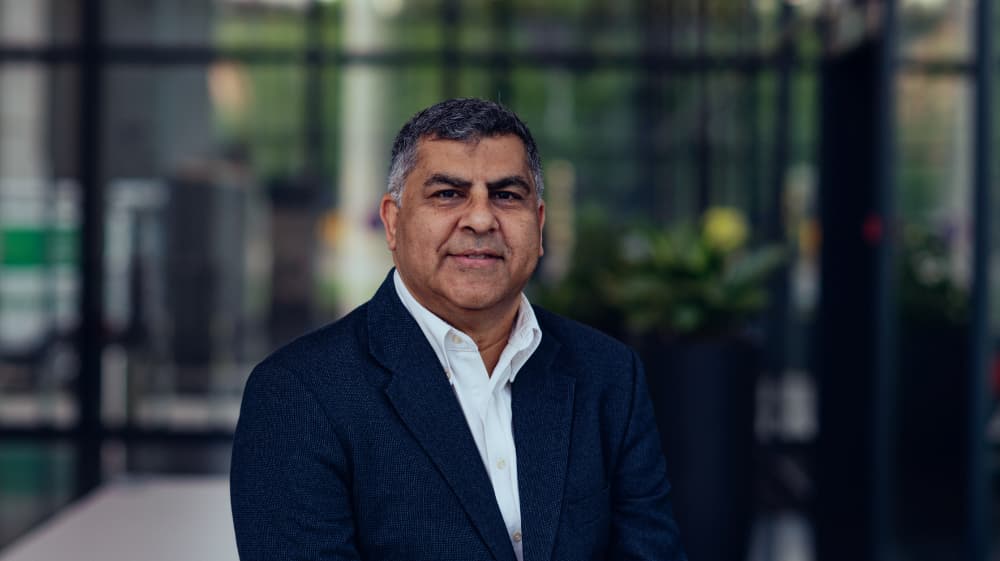 Vishal-Nayar Portrait of Vishal Nayar in a dark blazer and white shirt, standing in a modern glass-walled interior