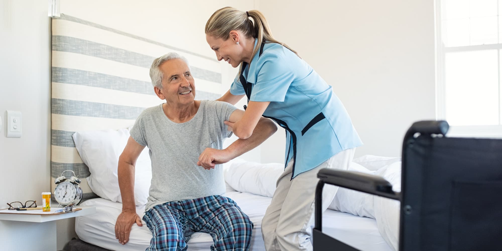 vital_sign_monitoring_header_1 Nurse helping an older man sit up on a bed in a bright hospital room