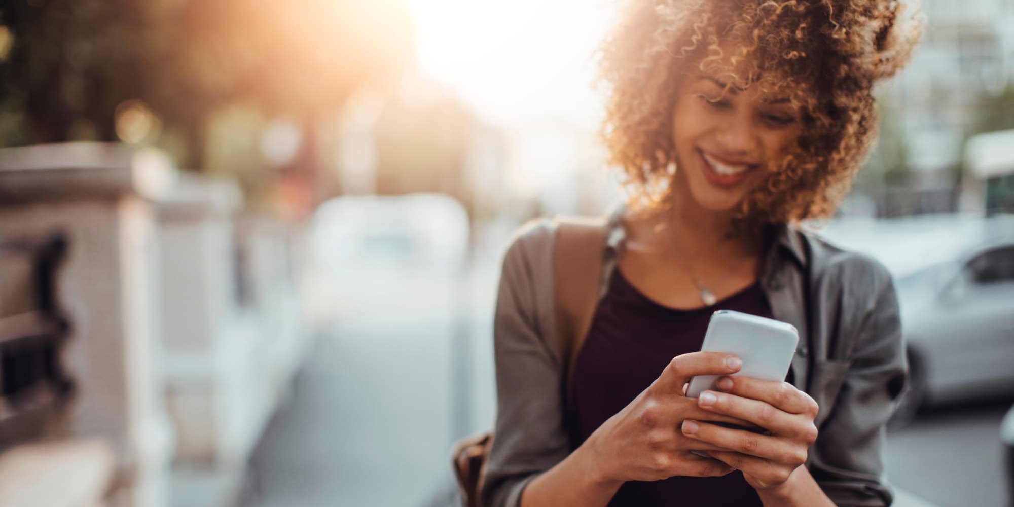 website_headers000x1000px_1 Smiling woman looking at a smartphone on a sunlit city street
