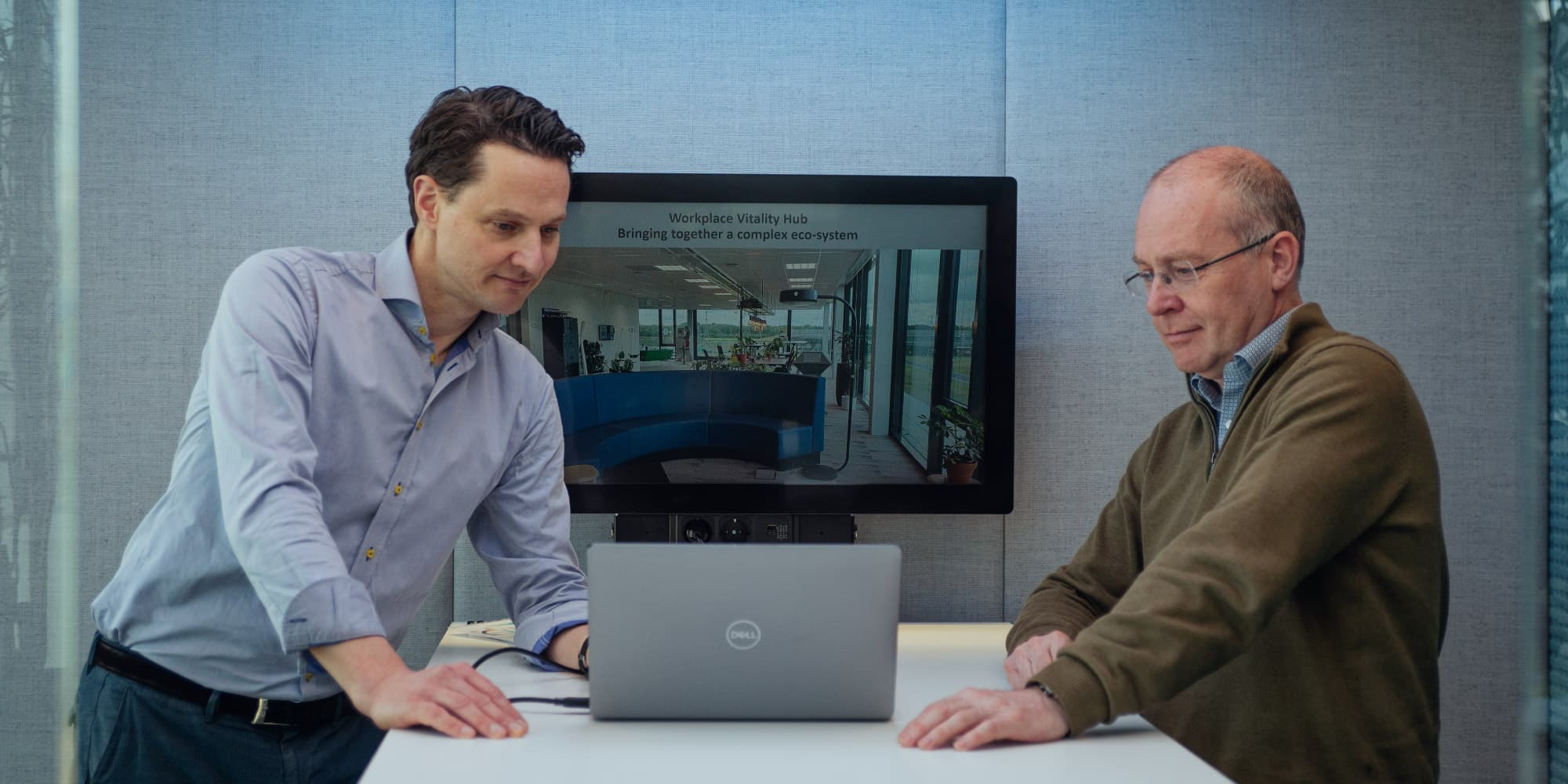 Where-innovation-meets-well-beingThe-Workplace-Vitality-Hub Two men discussing a laptop in an office, with a screen behind them showing the Workplace Vitality Hub.