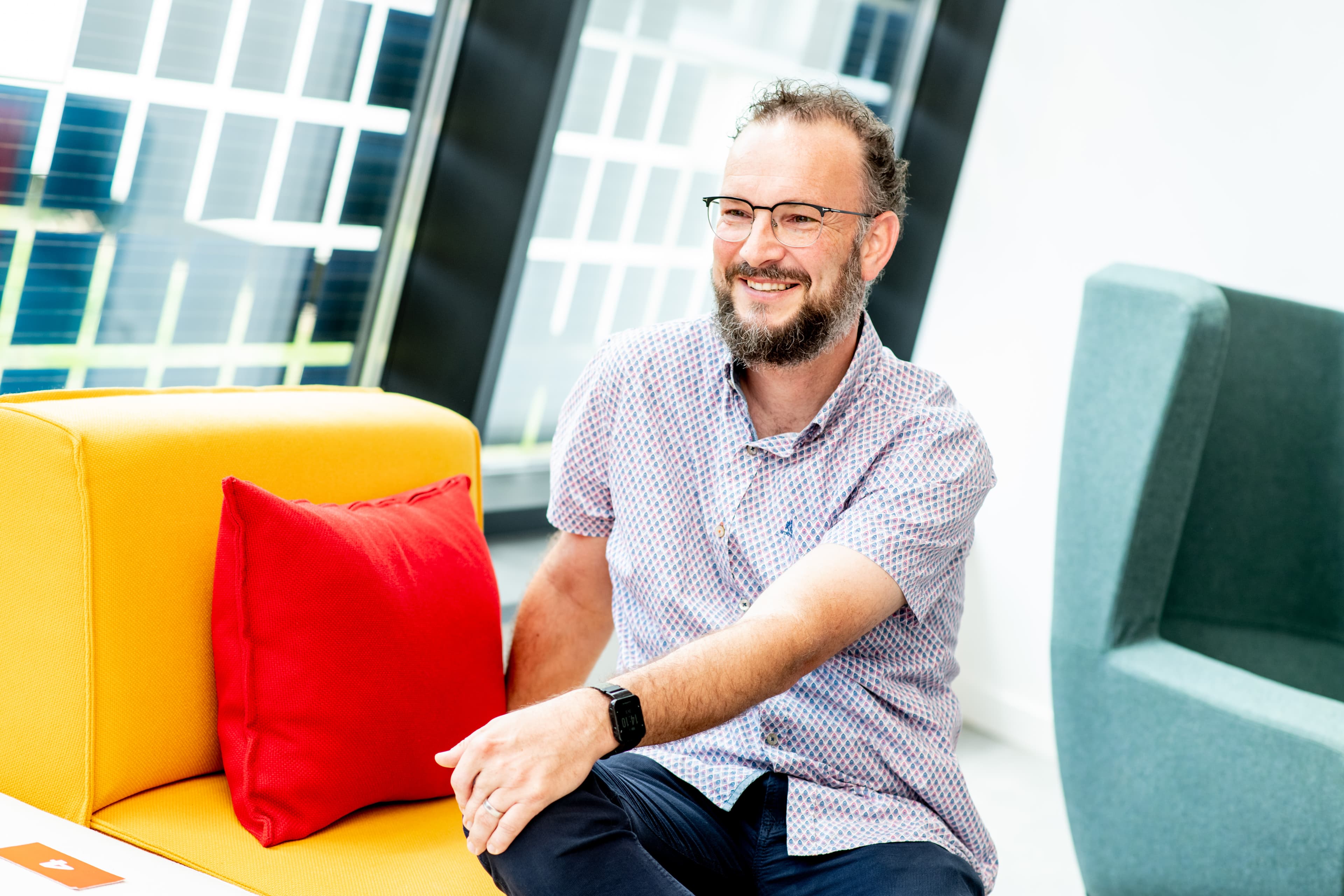 Wim%20Deferme-2 Wim Deferme smiling on a colorful lounge sofa with a red pillow in a bright office setting
