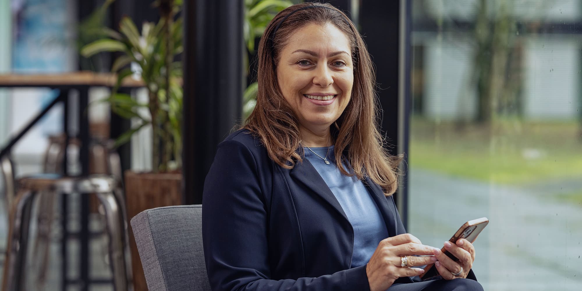 Womens-day1 Smiling woman in a navy blazer seated indoors, holding a smartphone by a large window.