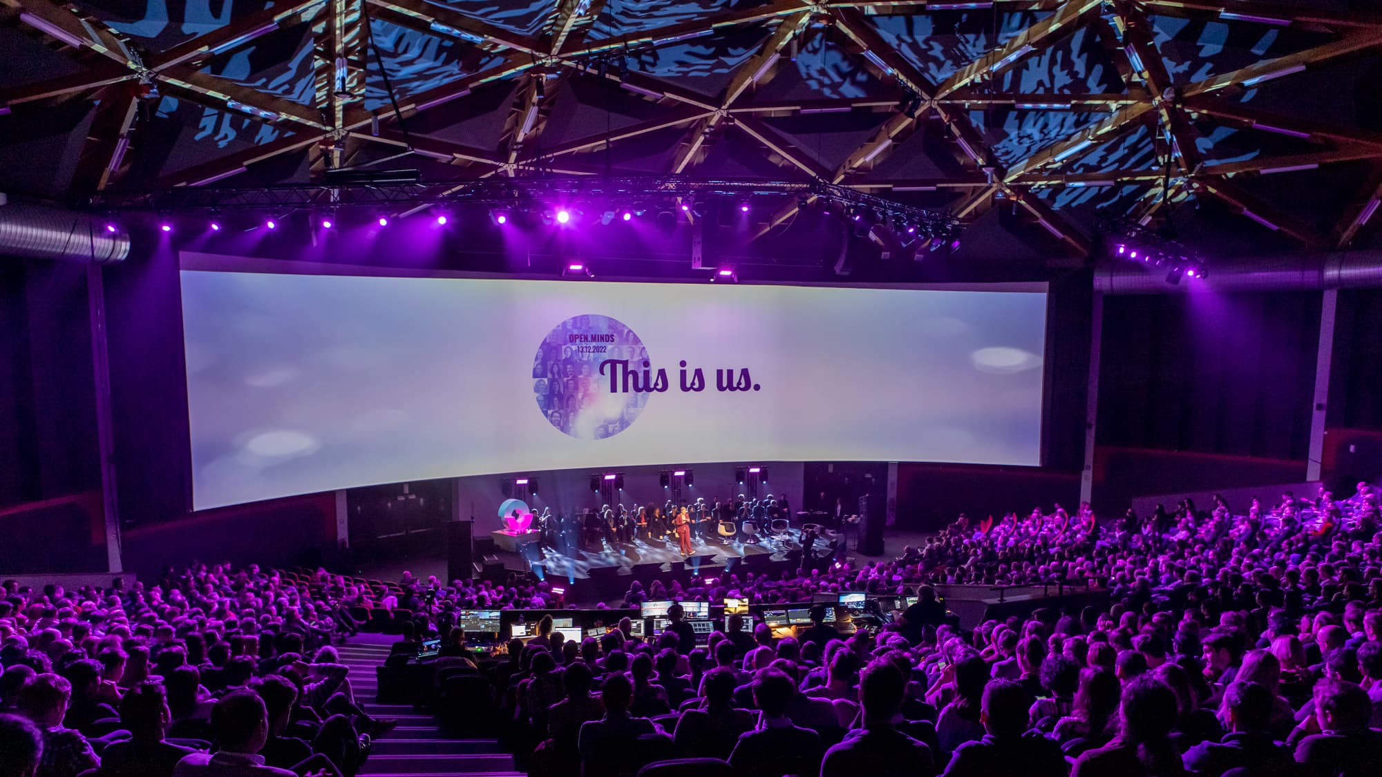 This-is-us Large auditorium with purple stage lighting, a screen reading “This is us.”, and an audience seated below
