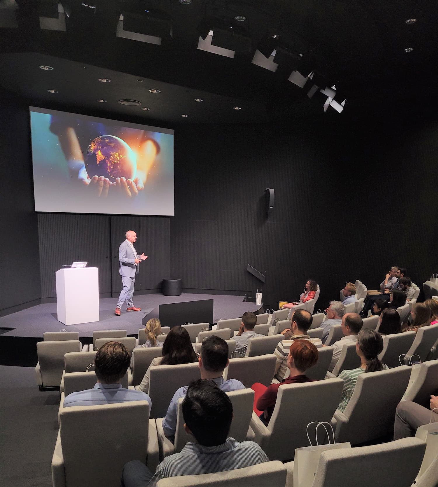 We-care-about-your-future Speaker presenting to an audience in an auditorium, with a slide showing hands holding a glowing Earth.