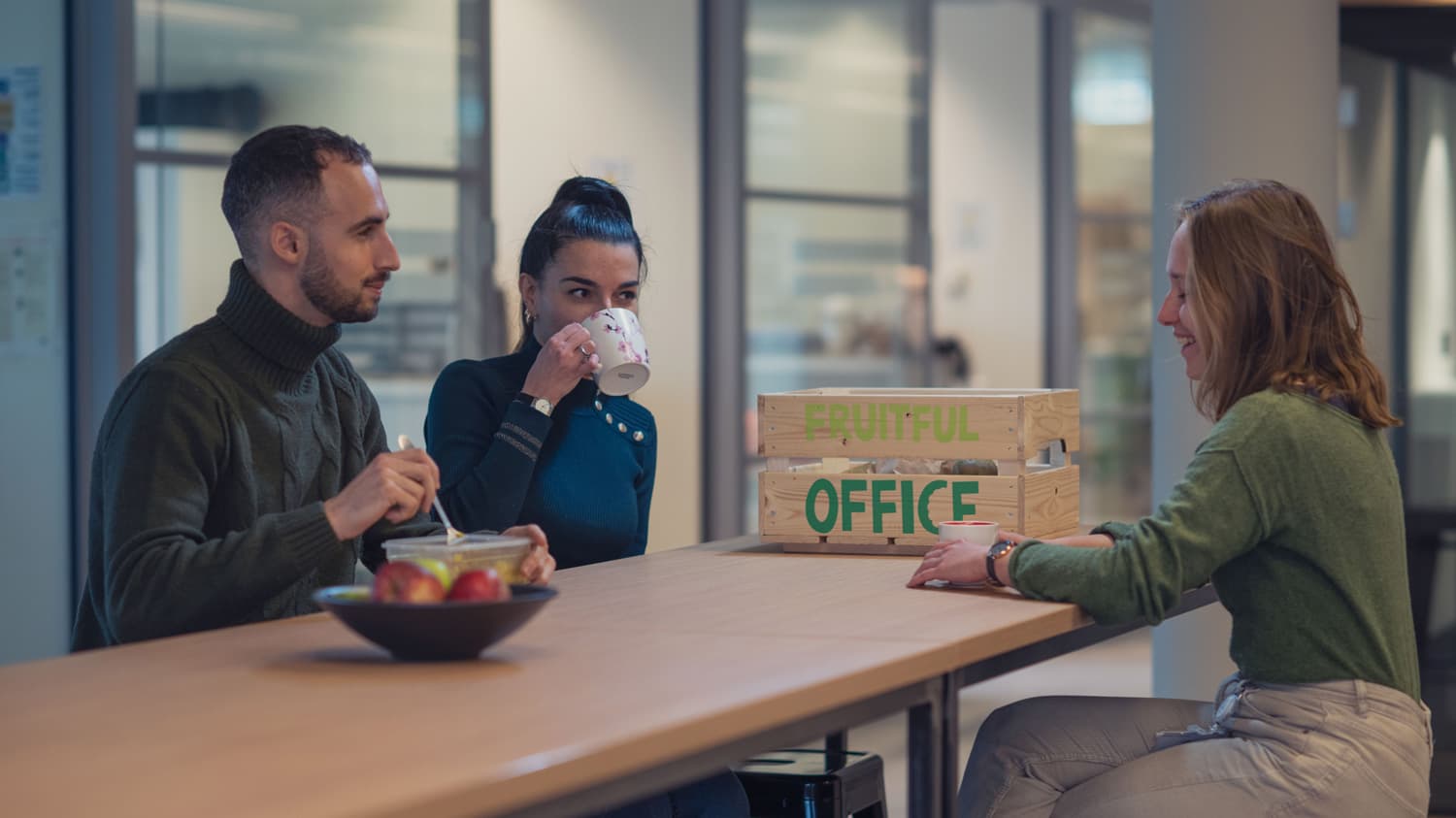 wellbeing Three coworkers chatting over coffee at a table with a fruit box labeled “FRUITFUL OFFICE”